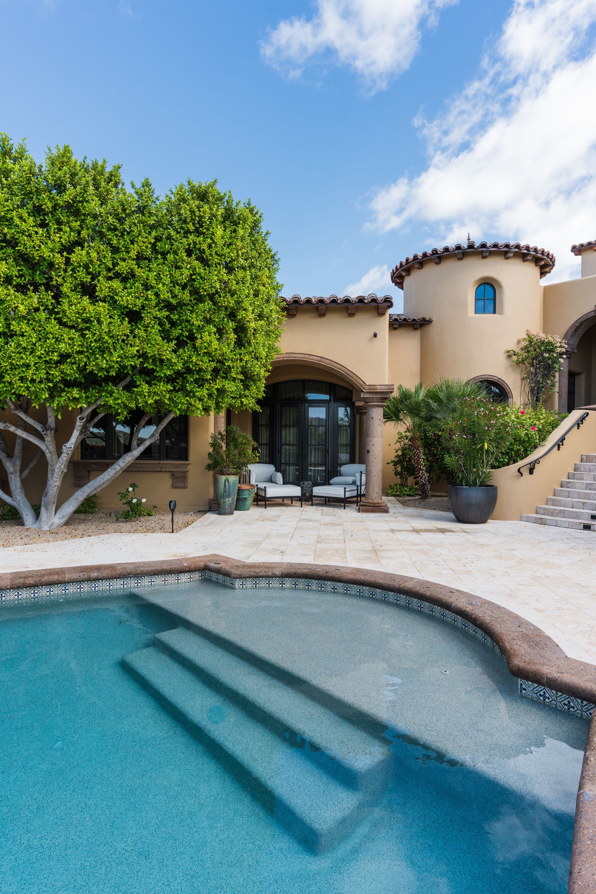 Pool with steps, leading to a beige stucco home with a curved tower and blue sky.