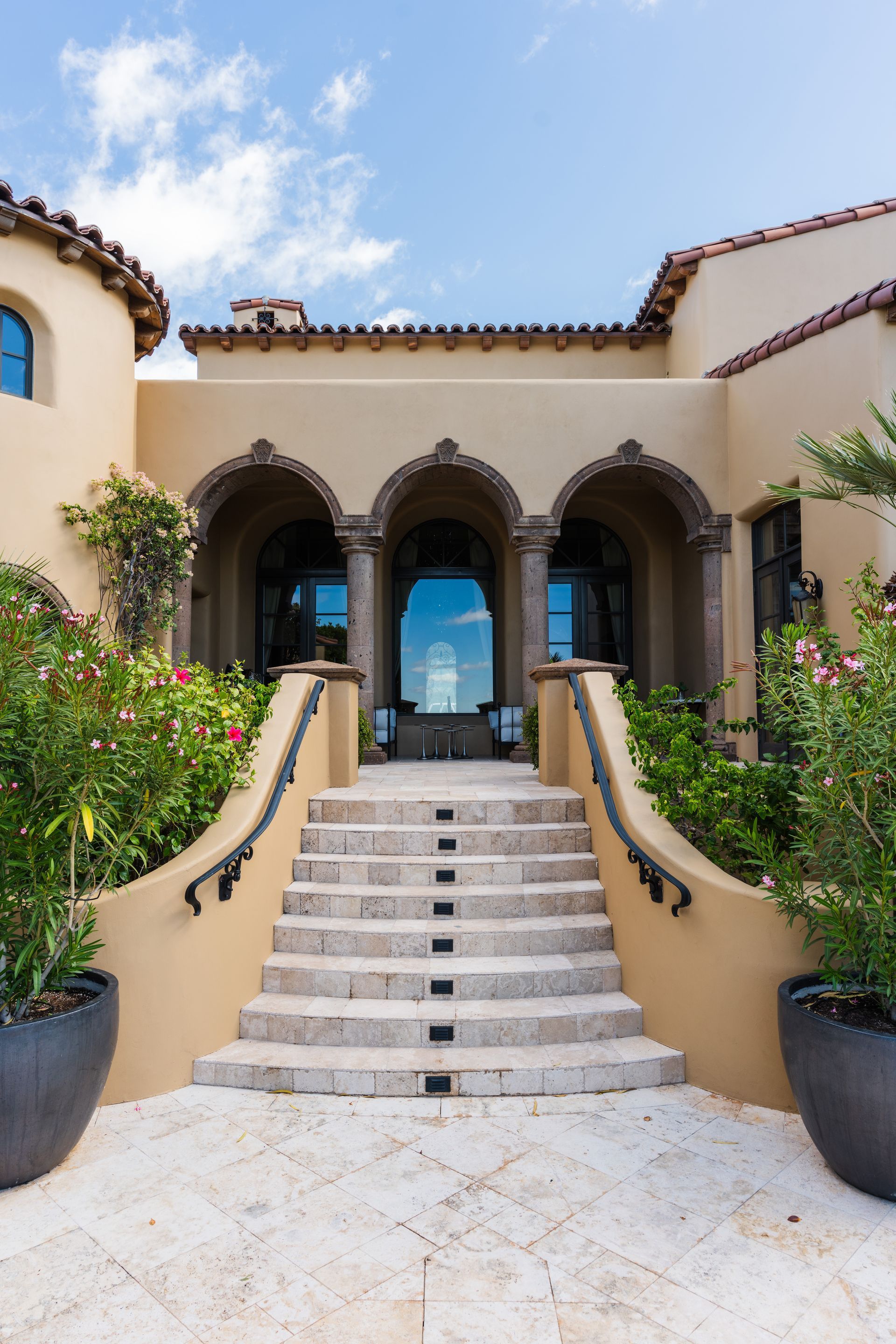 Stone steps leading up to an arched entrance with columns. Beige stucco exterior with lush landscaping and blue sky.