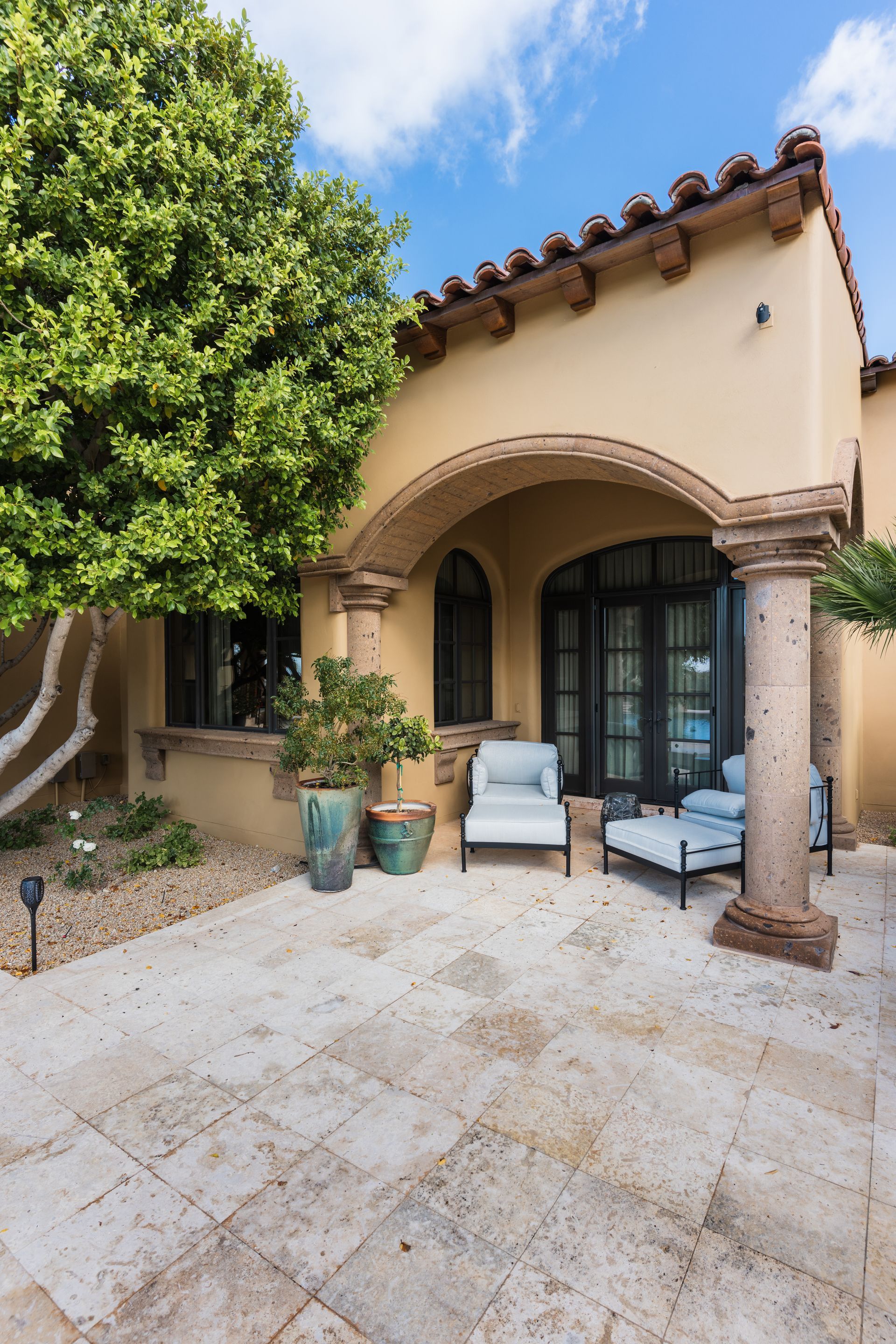 Patio with stone pavers, seating under arched entryway, beige stucco walls, and potted plants.