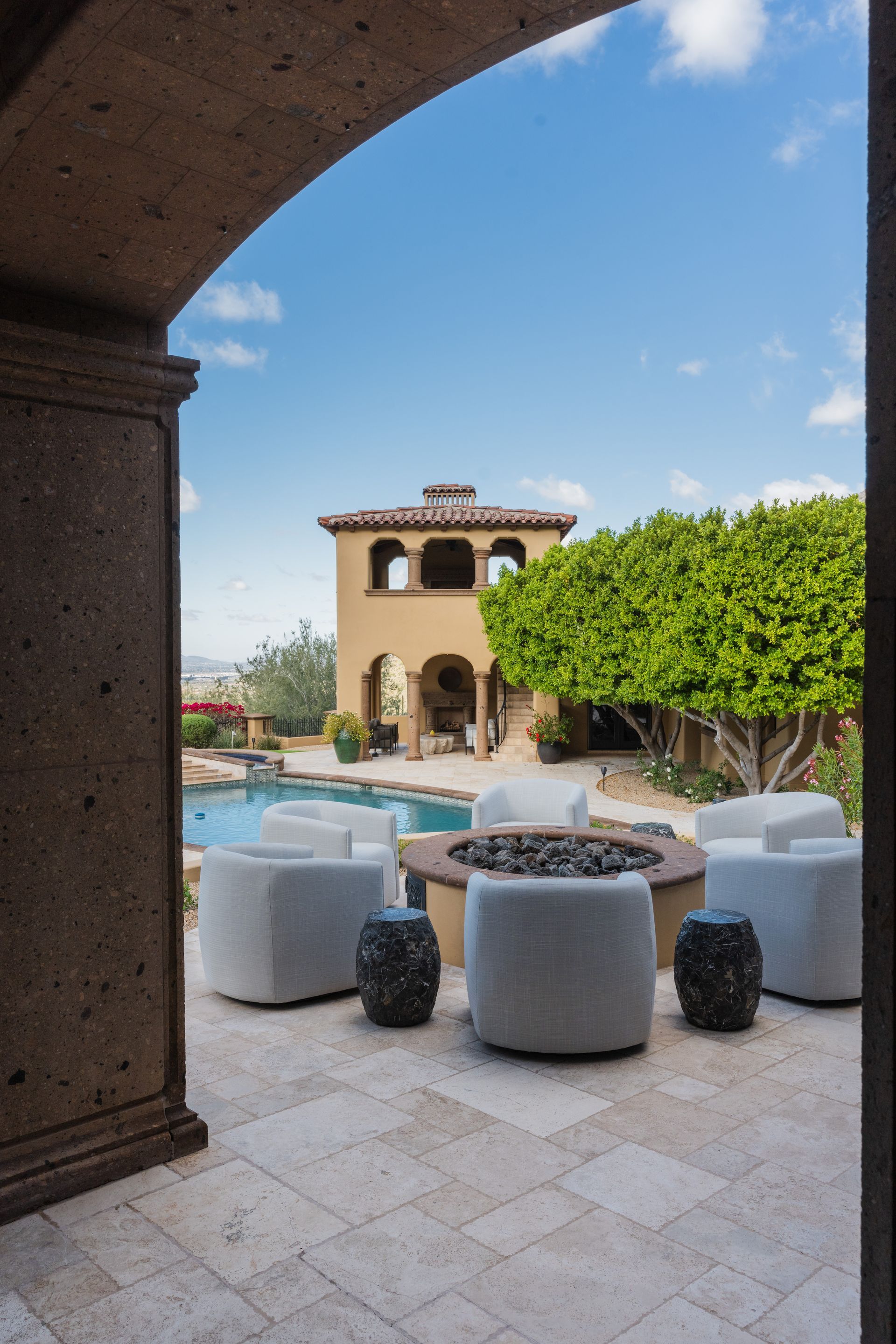 Patio with seating around a fire pit, pool, and tower in the distance, viewed through an arched doorway.