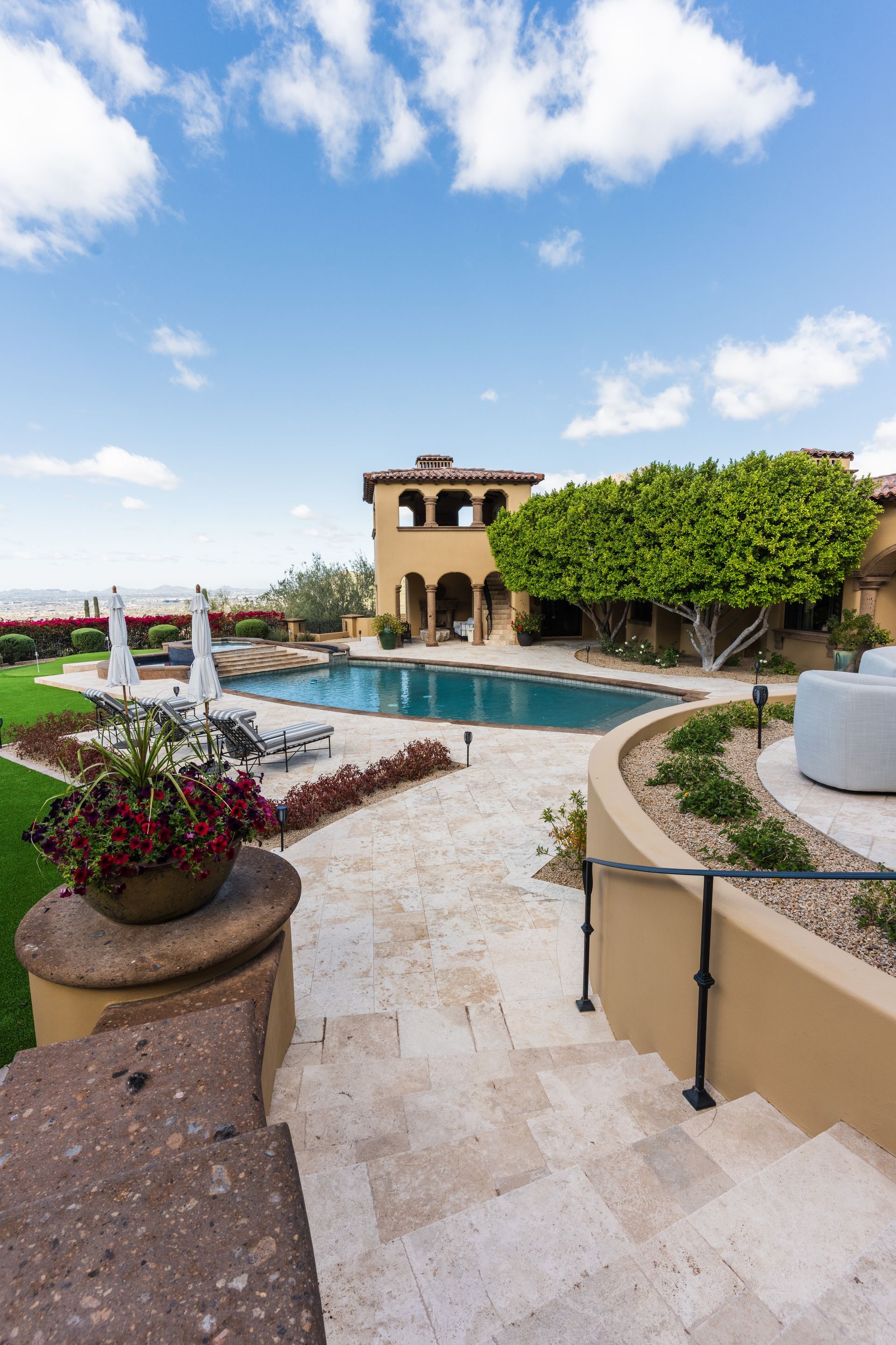 Exterior of a luxurious villa with a pool, stairs, patio, and lush landscaping on a sunny day.
