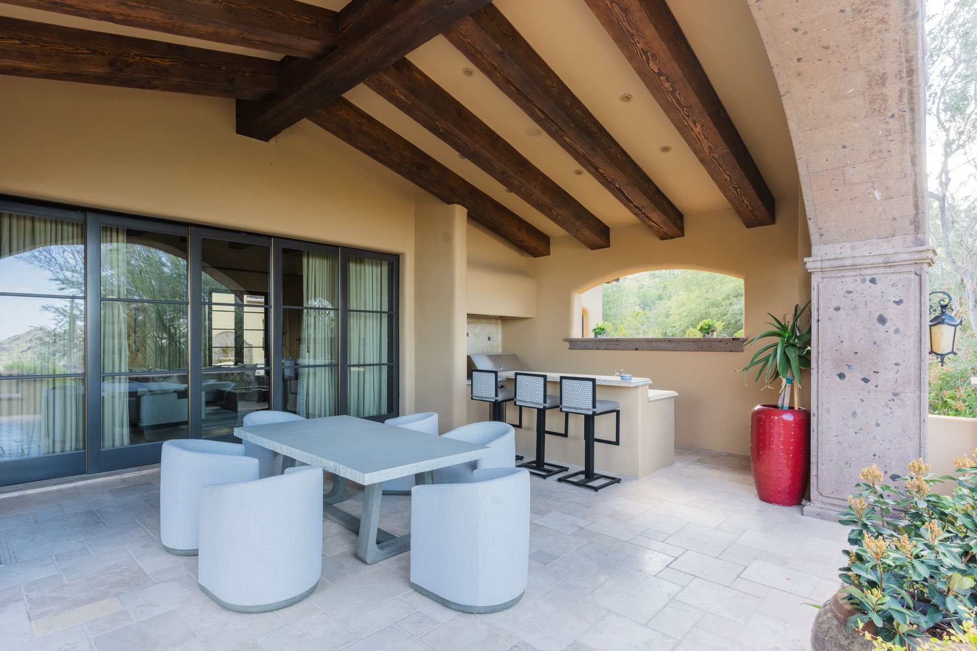 Patio with seating, outdoor kitchen, and dark wood beams. Beige walls, stone pillars, and sliding glass doors.