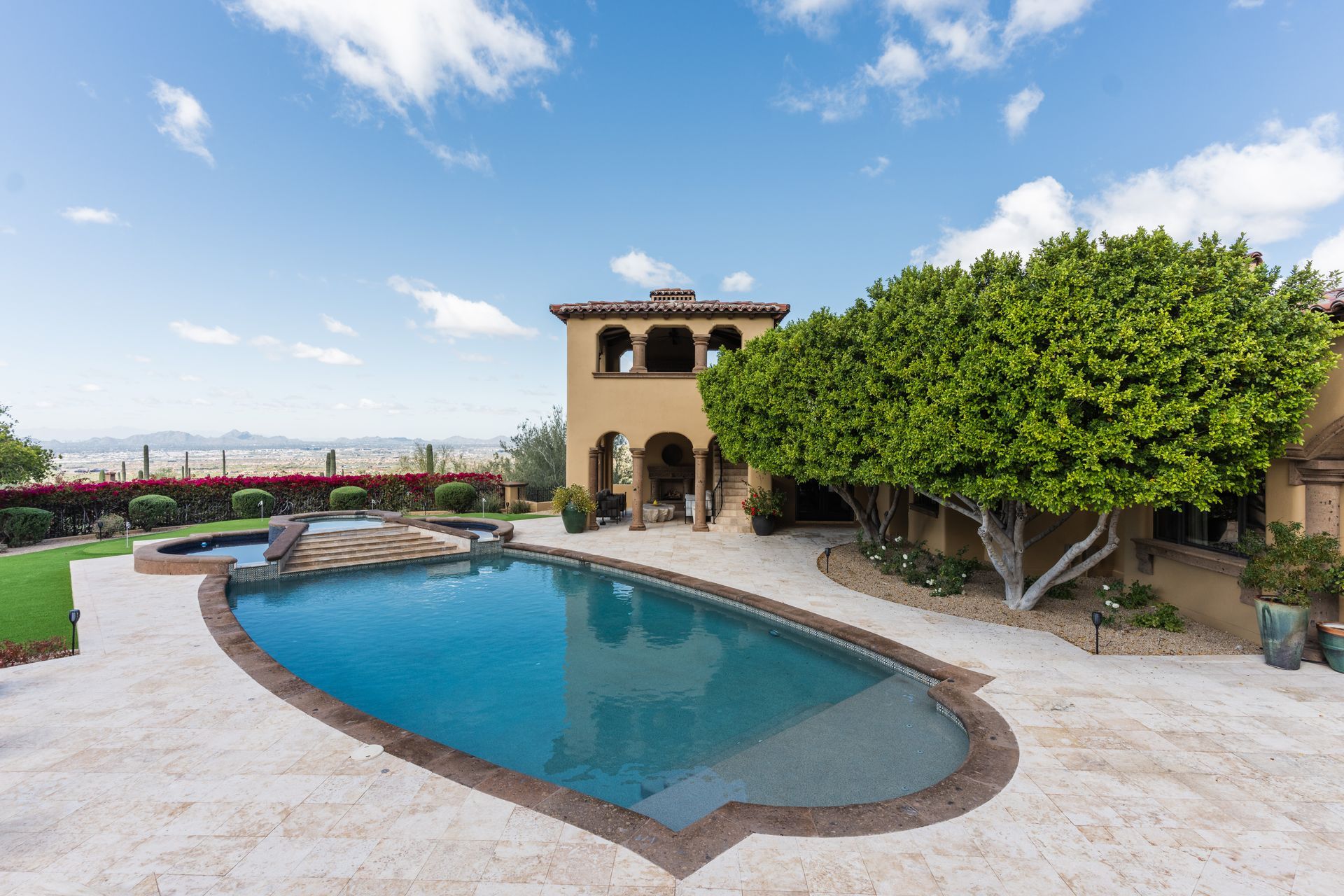 Swimming pool in front of a tan two-story house with arches, blue sky, and a lush green tree.