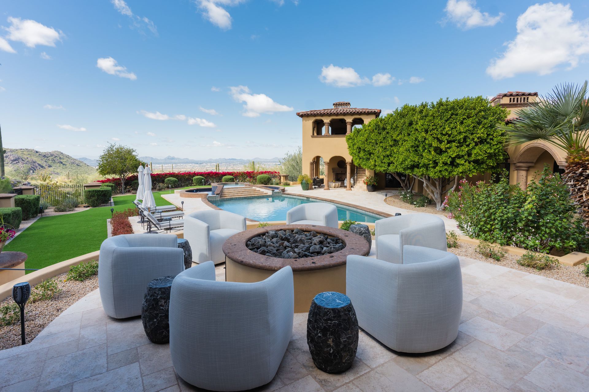 Patio with fire pit, seating, pool, and house under a blue sky.