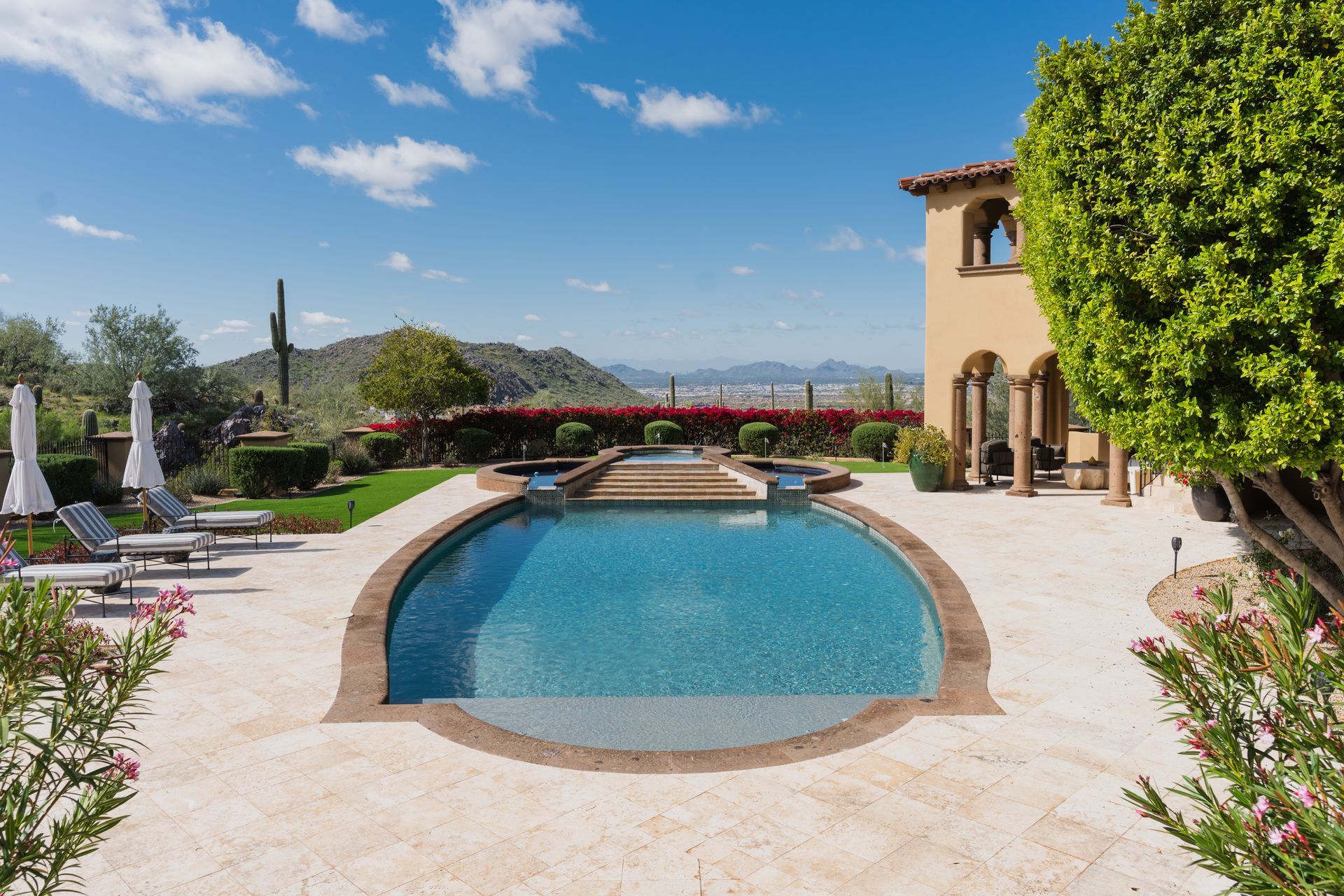 Backyard pool with mountain views, sunny day, beige patio, lush greenery, and a two-story beige house.