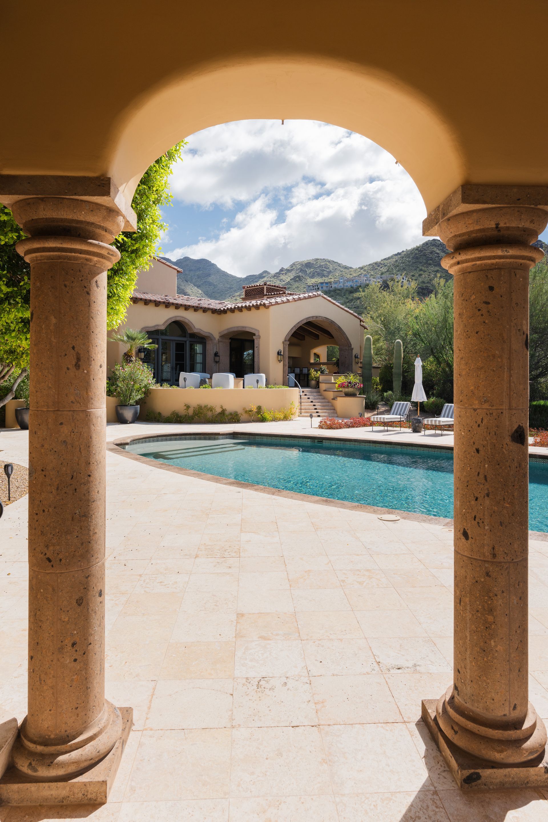 Stone columns frame a view of a house, pool, and mountains under a cloudy sky.