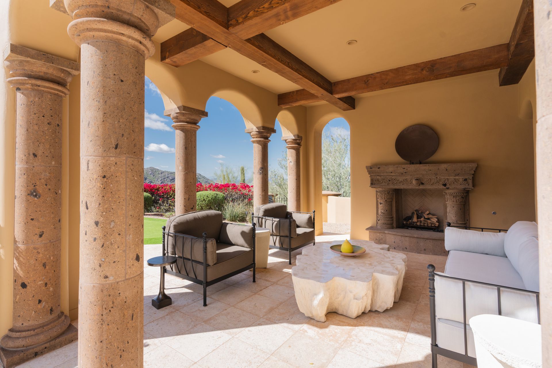 Outdoor patio with stone columns, fireplace, seating, and a view of a garden under a blue sky.