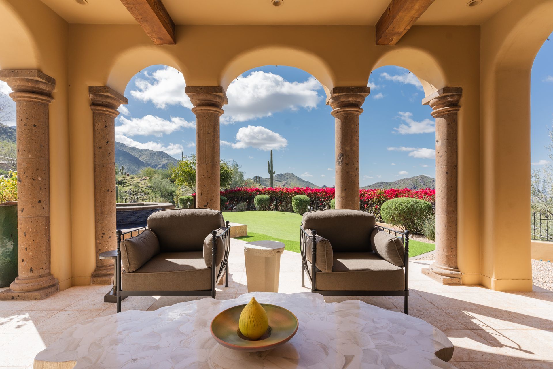 Outdoor patio with arched columns, two chairs, and a table overlooking a garden and mountains.