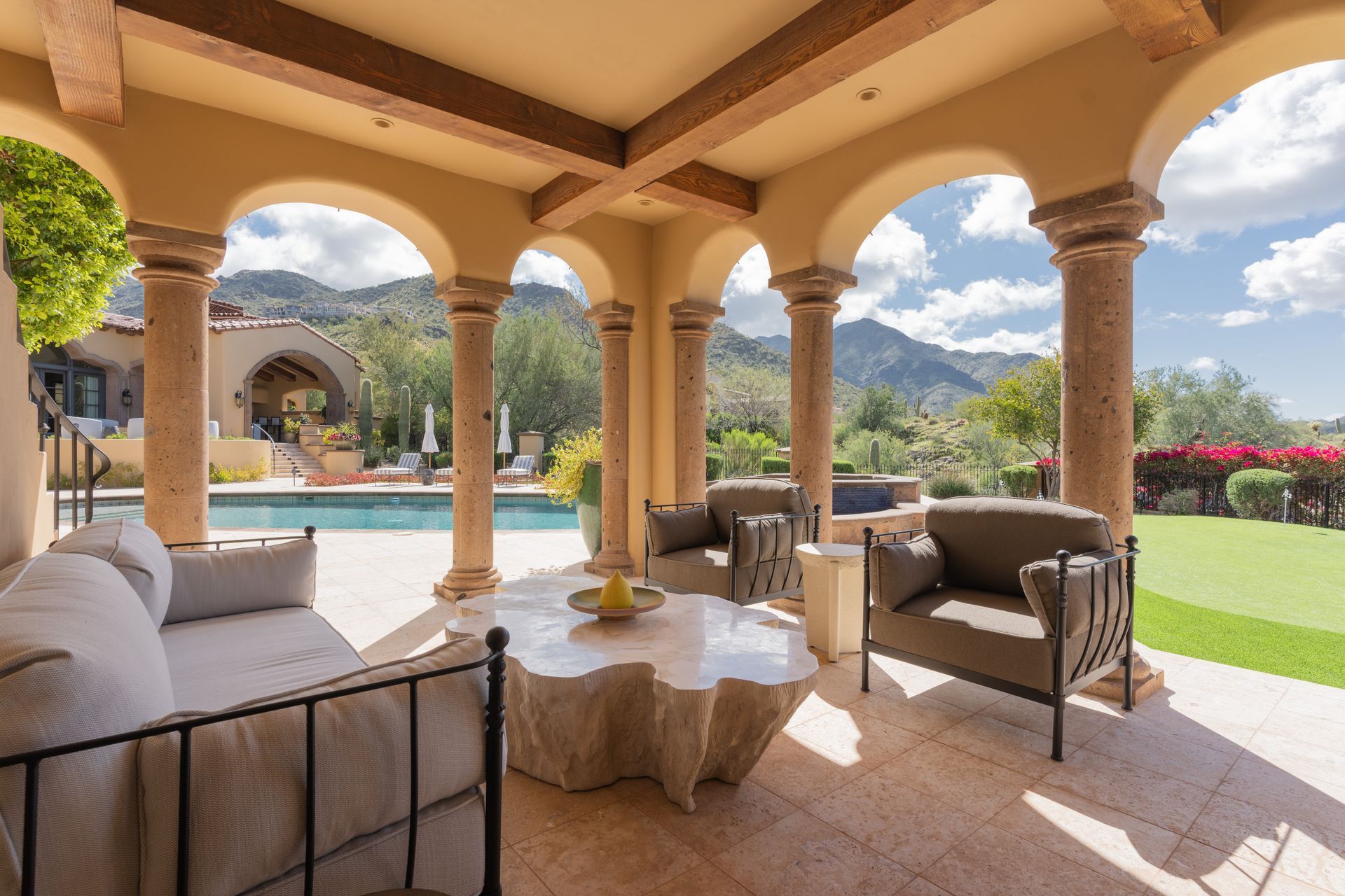 Covered patio with seating overlooking a pool, mountains, and sky. Beige columns and stone table.