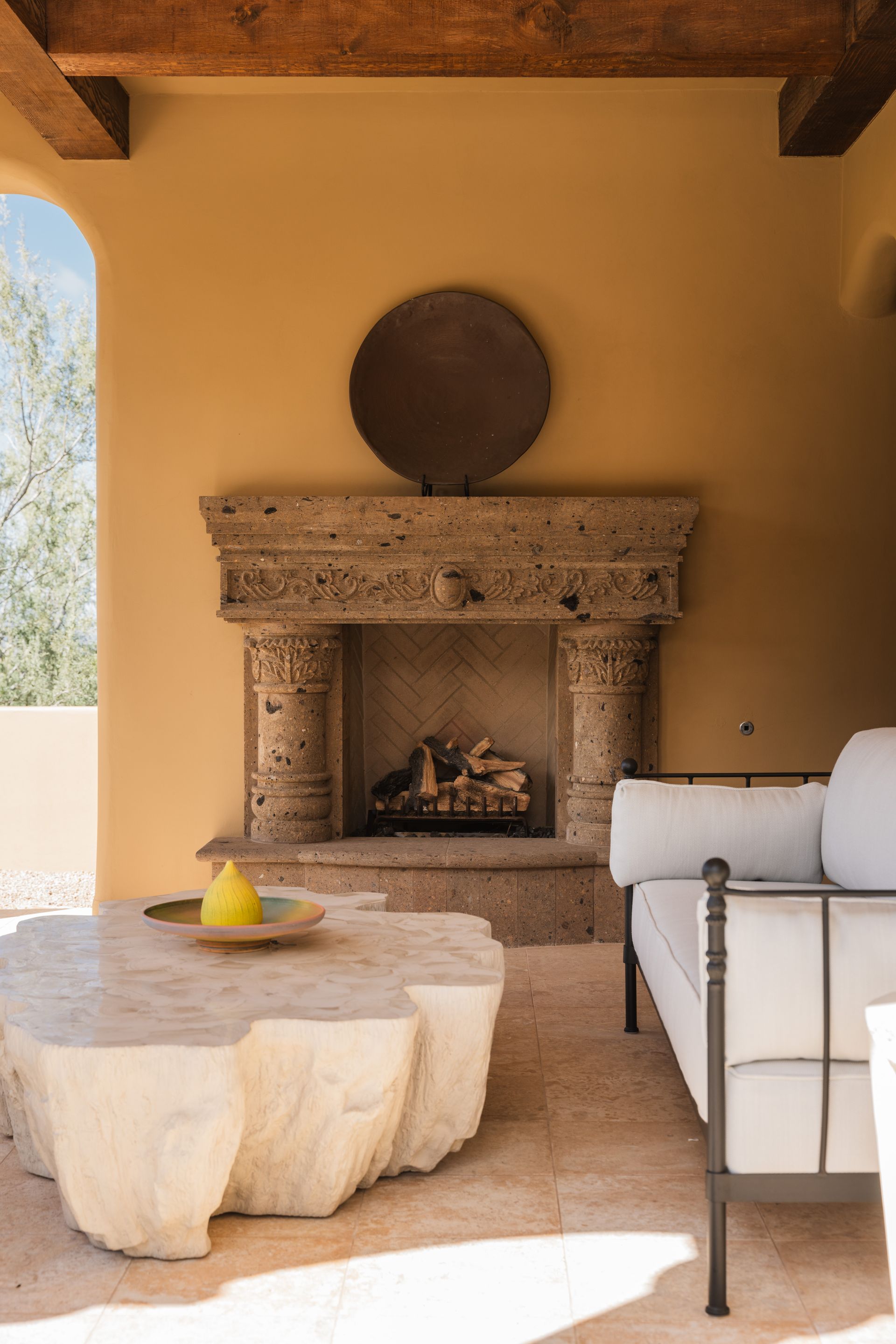 Outdoor living space with stone fireplace, coffee table, and couch. Beige walls, wood beams, and a round metal art piece.