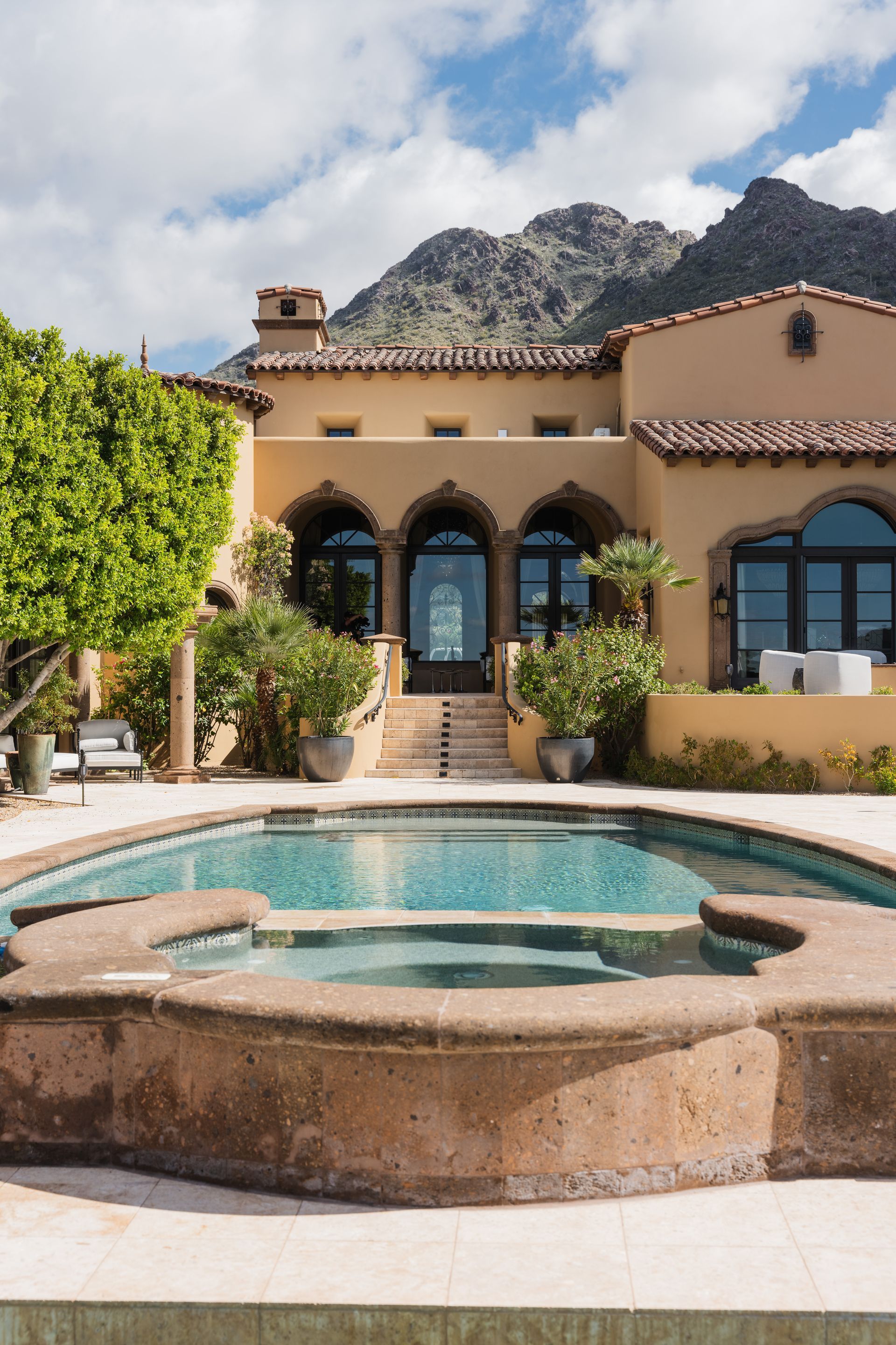 Swimming pool in front of a tan stucco home with arched windows, mountains in the background.