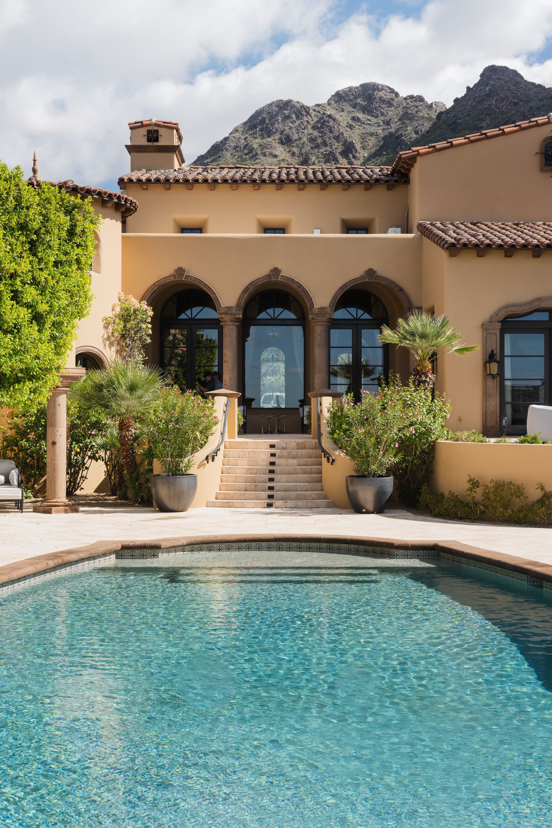 Luxury home with pool and mountain backdrop; arched doorways, tile roof, and turquoise water.