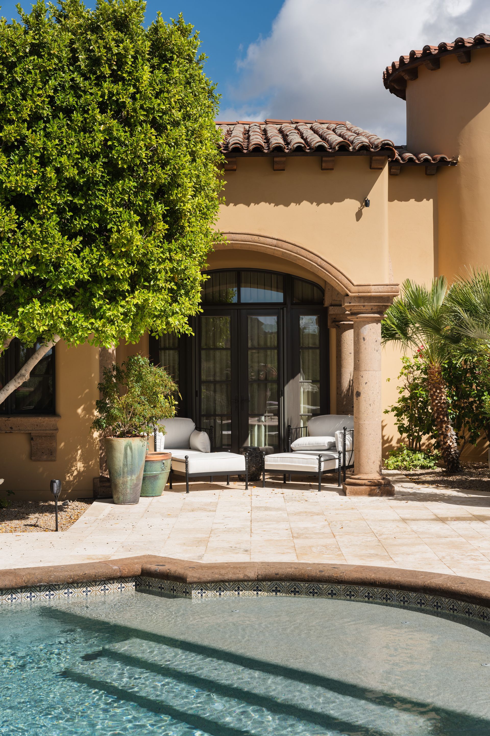 Backyard patio with pool, lounge chairs, and arched doorway, under sunny skies.