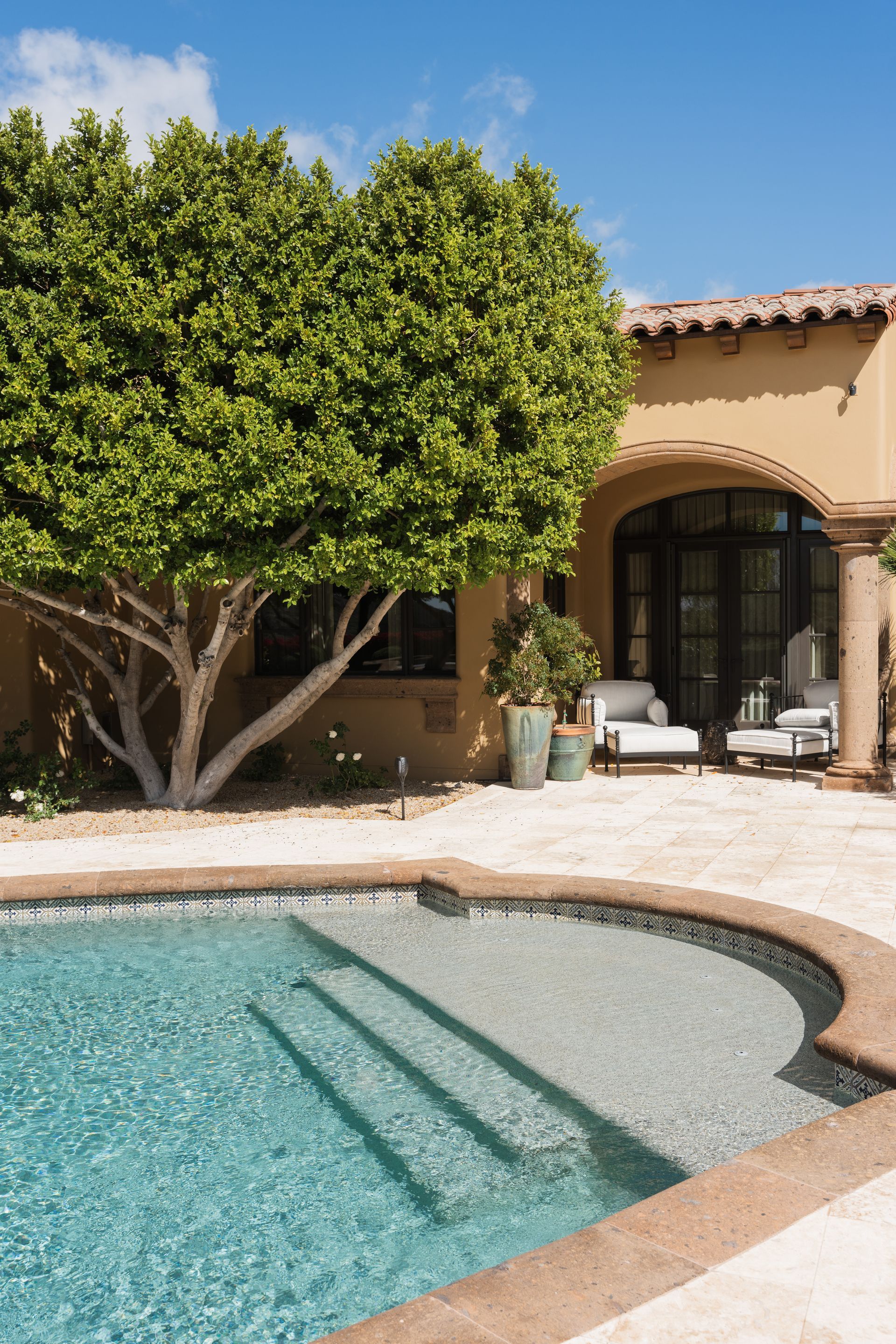 Pool with steps, next to a tree and patio with seating, under a sunny sky.