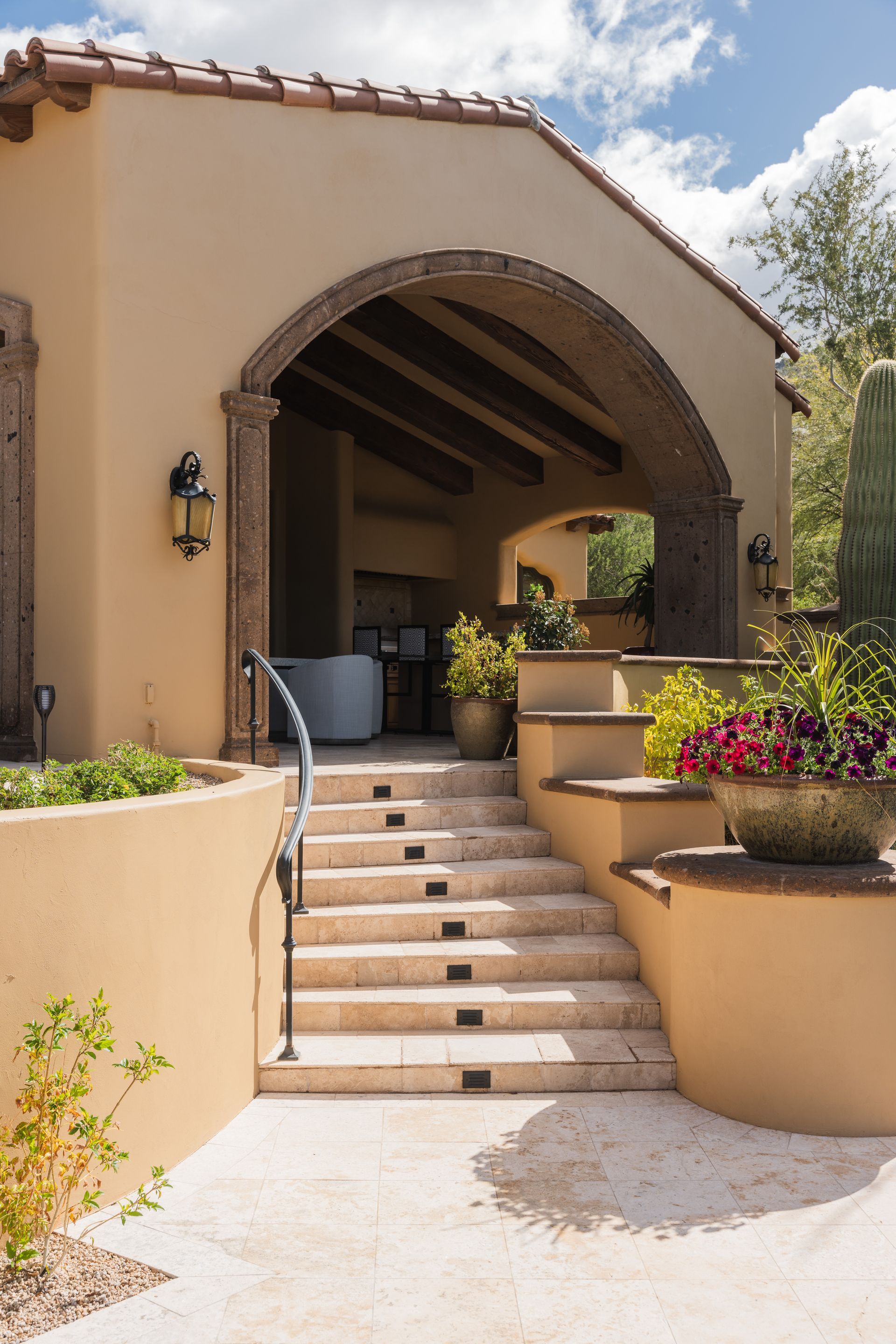 Outdoor steps leading up to a covered patio with an arched entryway. Beige stucco, terracotta roof tiles, and potted plants.