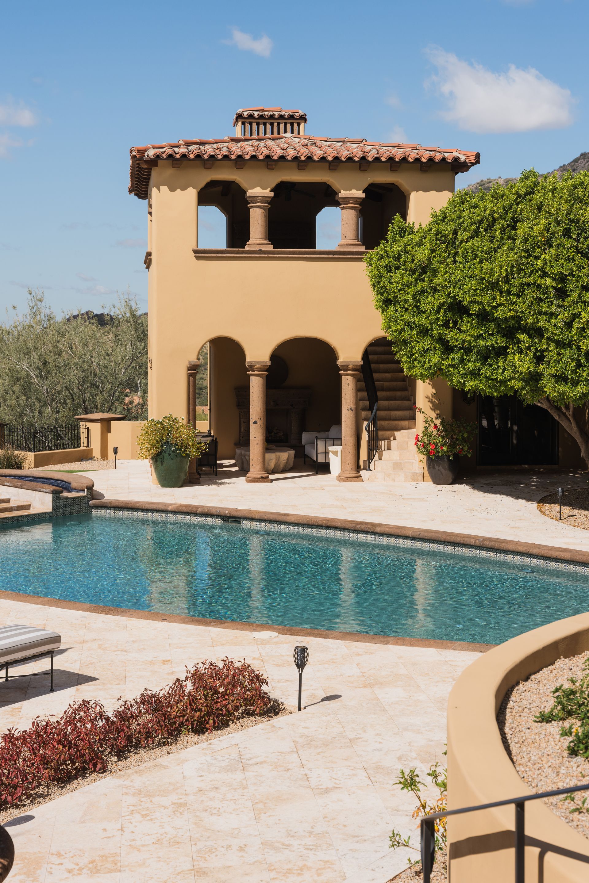 Beige tower with arched openings beside a pool. Brown tile roof, stone patio, green plants, and blue water.