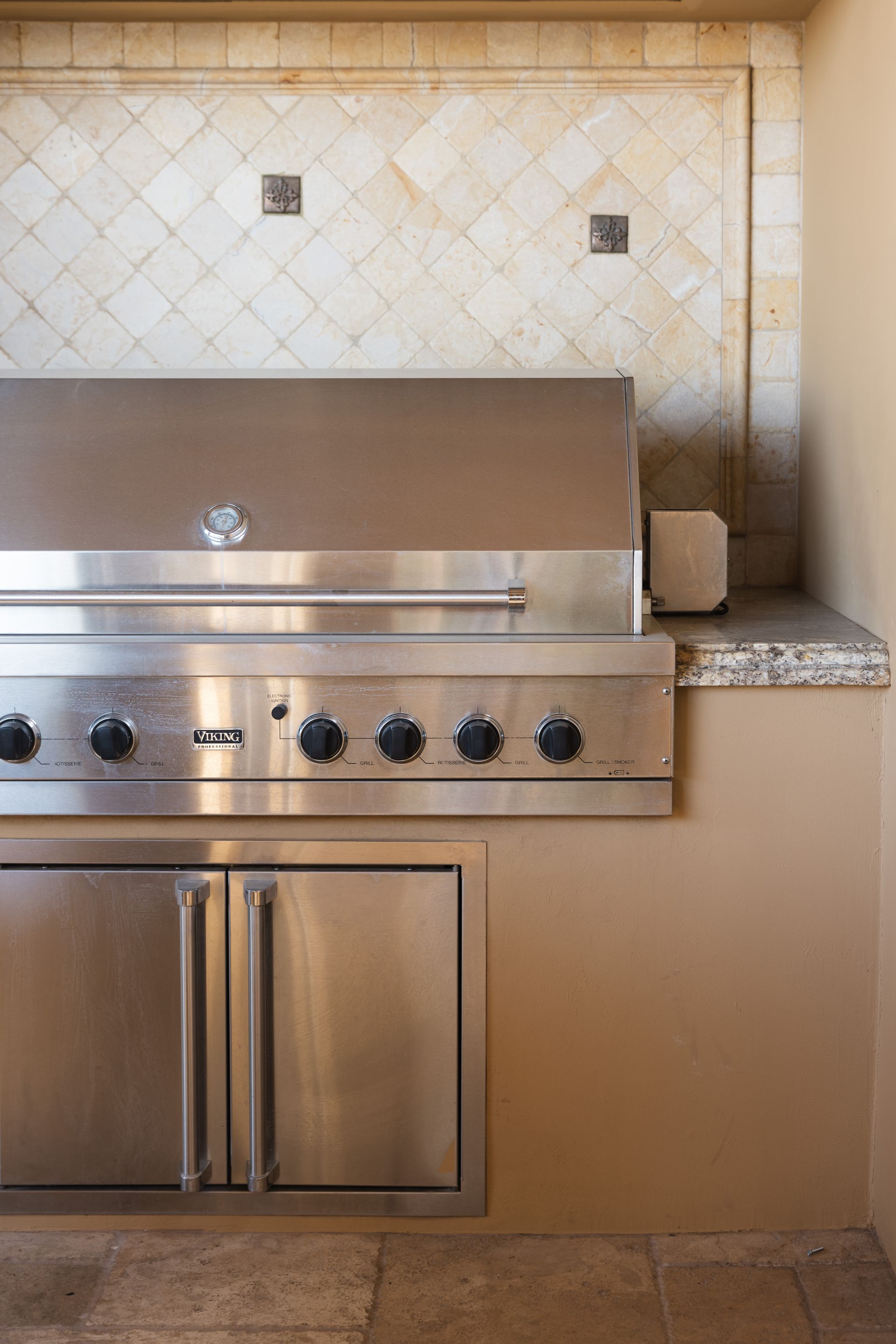 Stainless steel outdoor grill and refrigerator set into a beige stucco wall with stone tile backsplash.