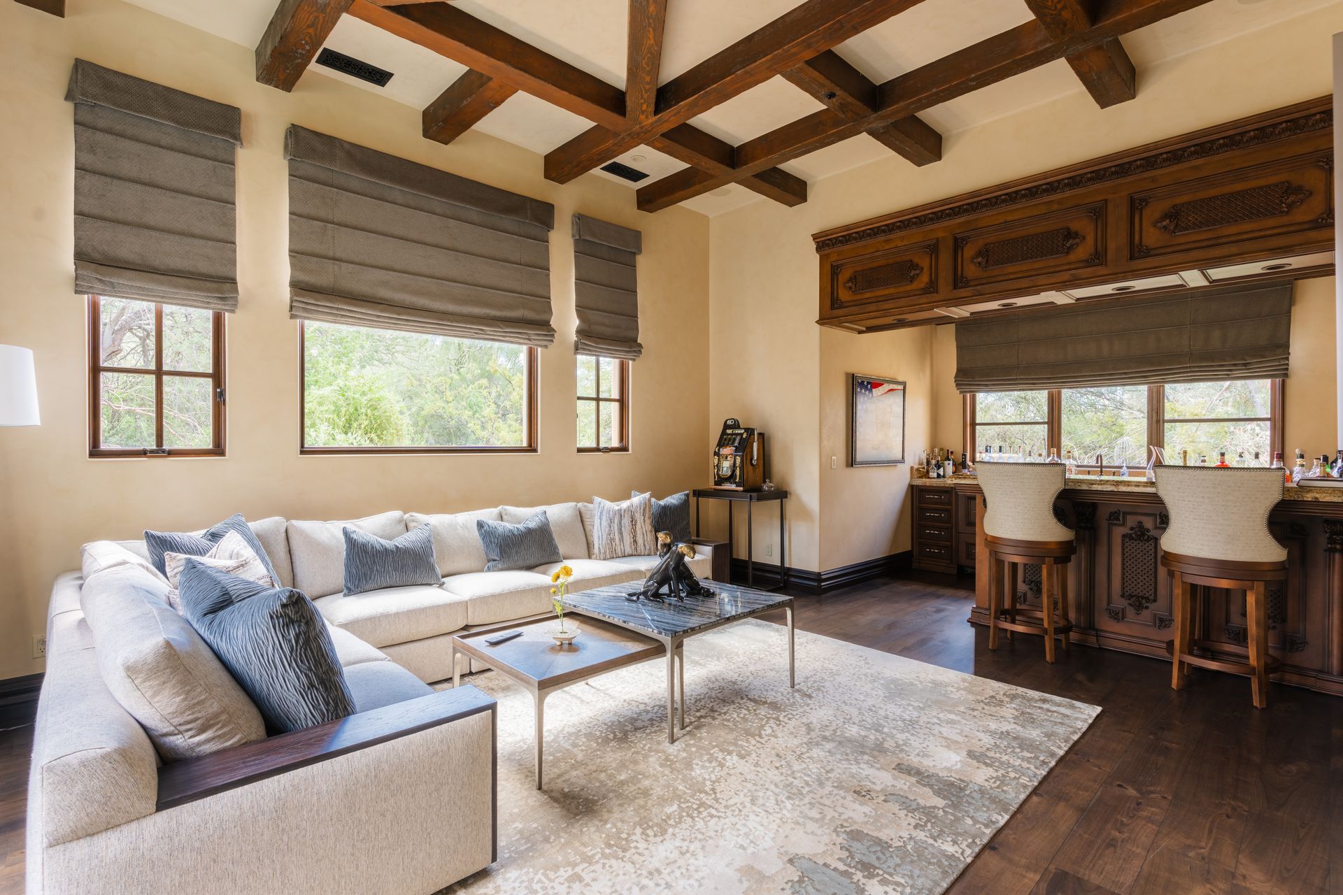 Living room with a large sectional sofa, rug, and bar area; featuring wooden beams and Roman shades.