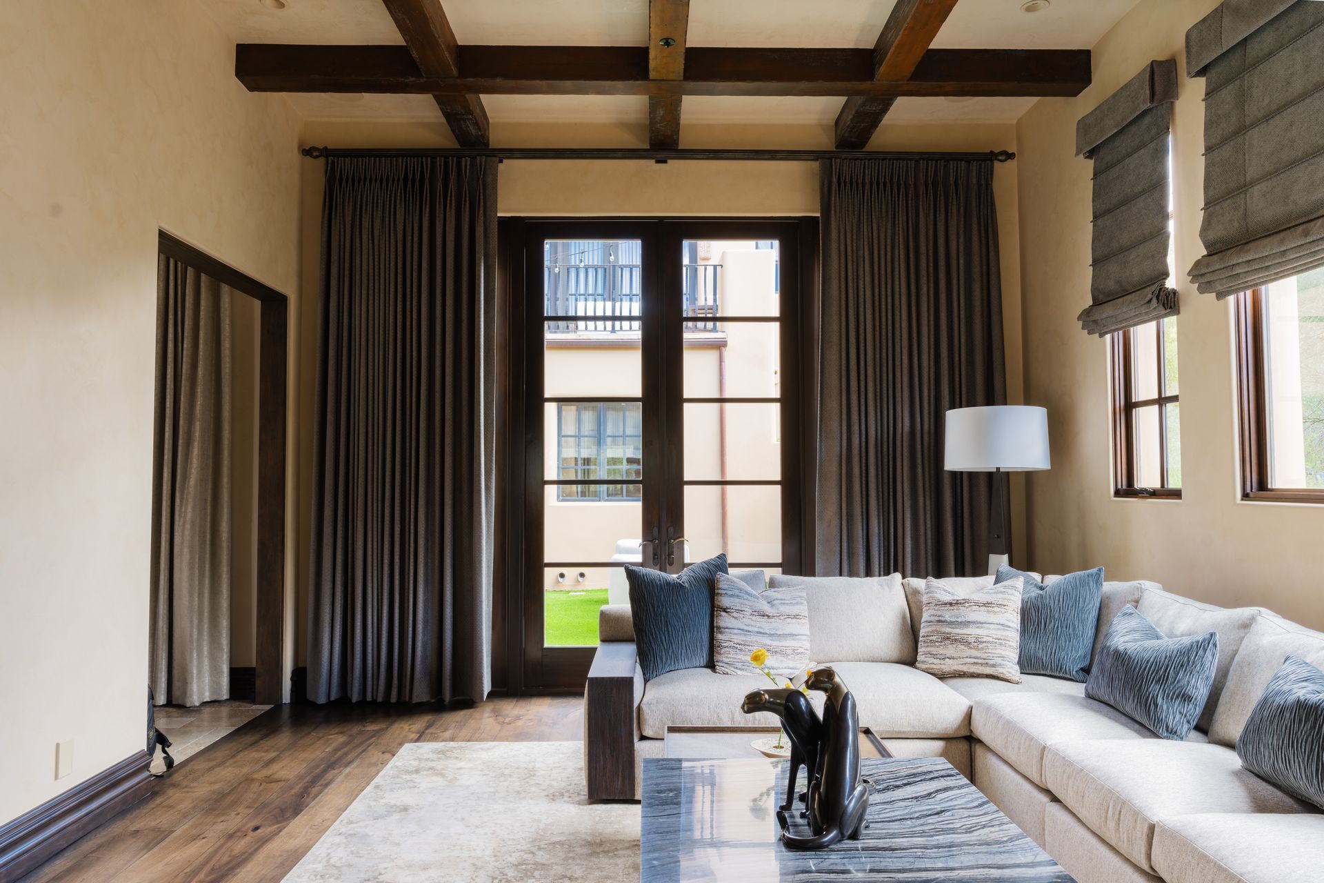 Living room with cream-colored sectional sofa, large windows with gray curtains, wooden beams, and a beige wall.