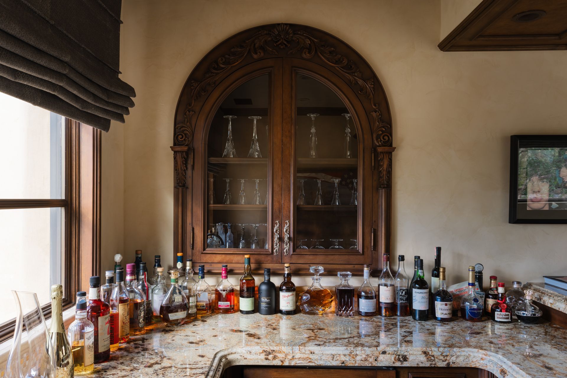 Bar area with ornate wooden cabinet and liquor bottles on a granite countertop. Window with shade on the left.