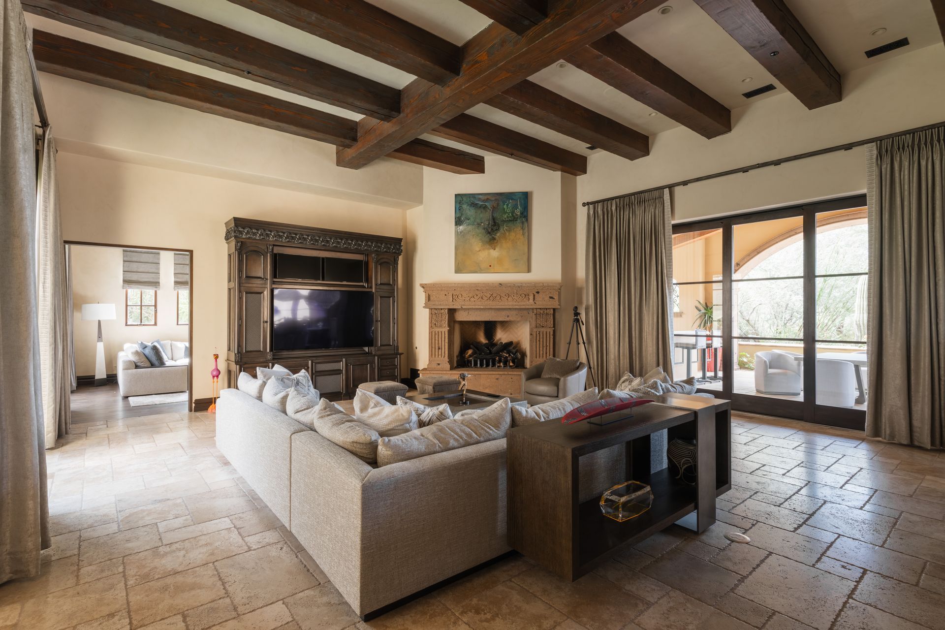 Living room with beige walls, beamed ceiling, fireplace, large TV, sofa, and glass doors to a patio.