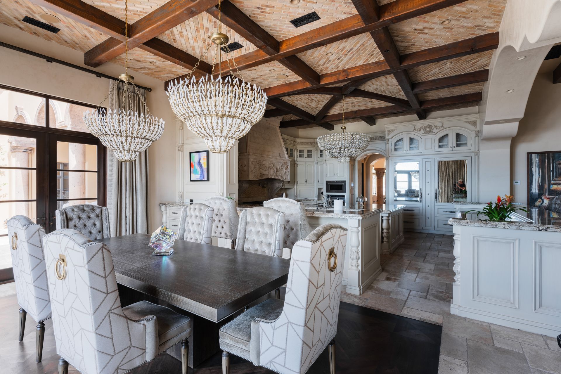 Dining room with large chandelier, ornate chairs, dark wood beams, and a white kitchen in the background.