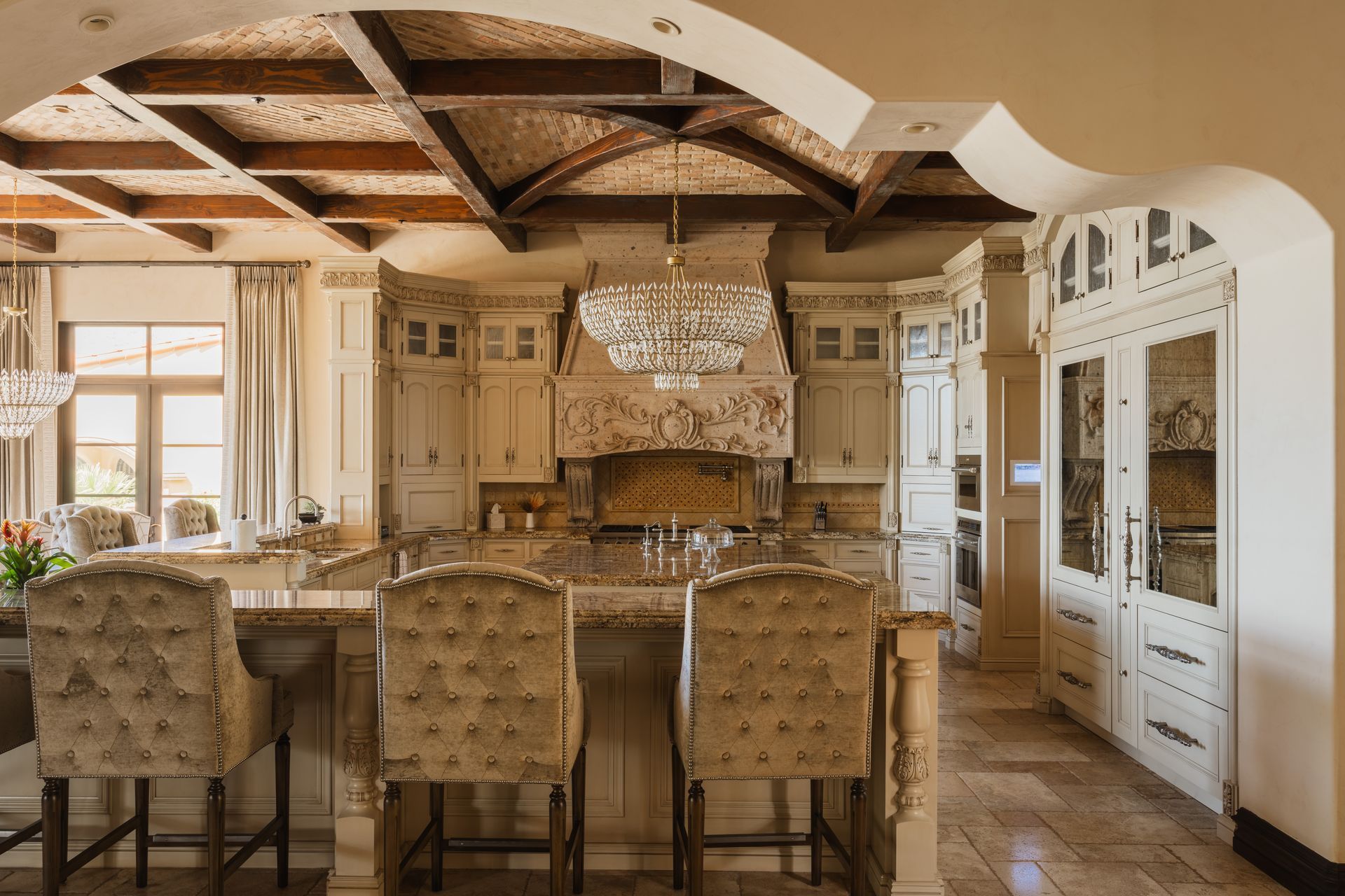 Elegant kitchen with cream cabinets, decorative range hood, island with stools, and tiled floor.