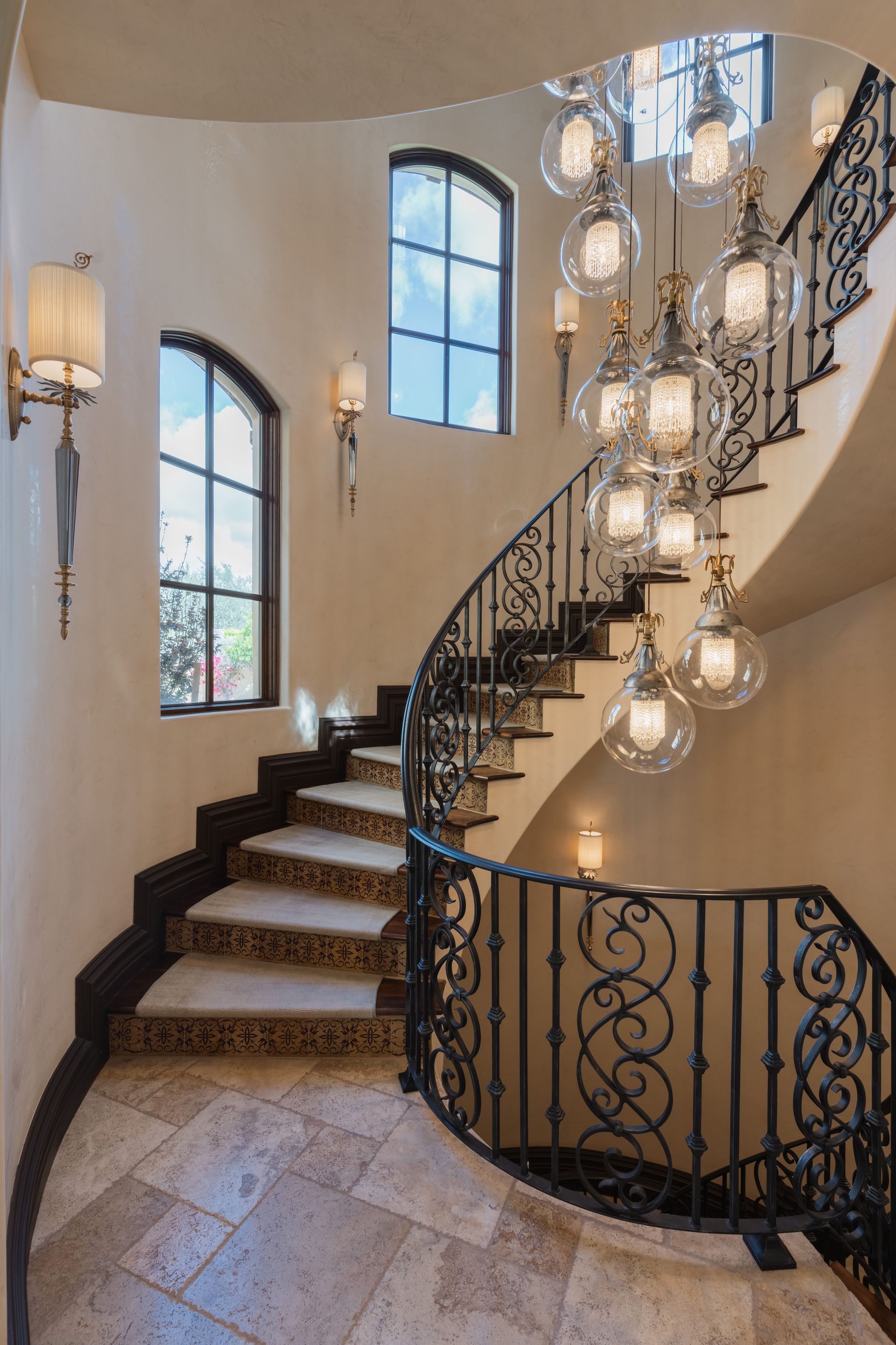 Elegant curved staircase with wrought iron railing, chandelier, and arched windows.