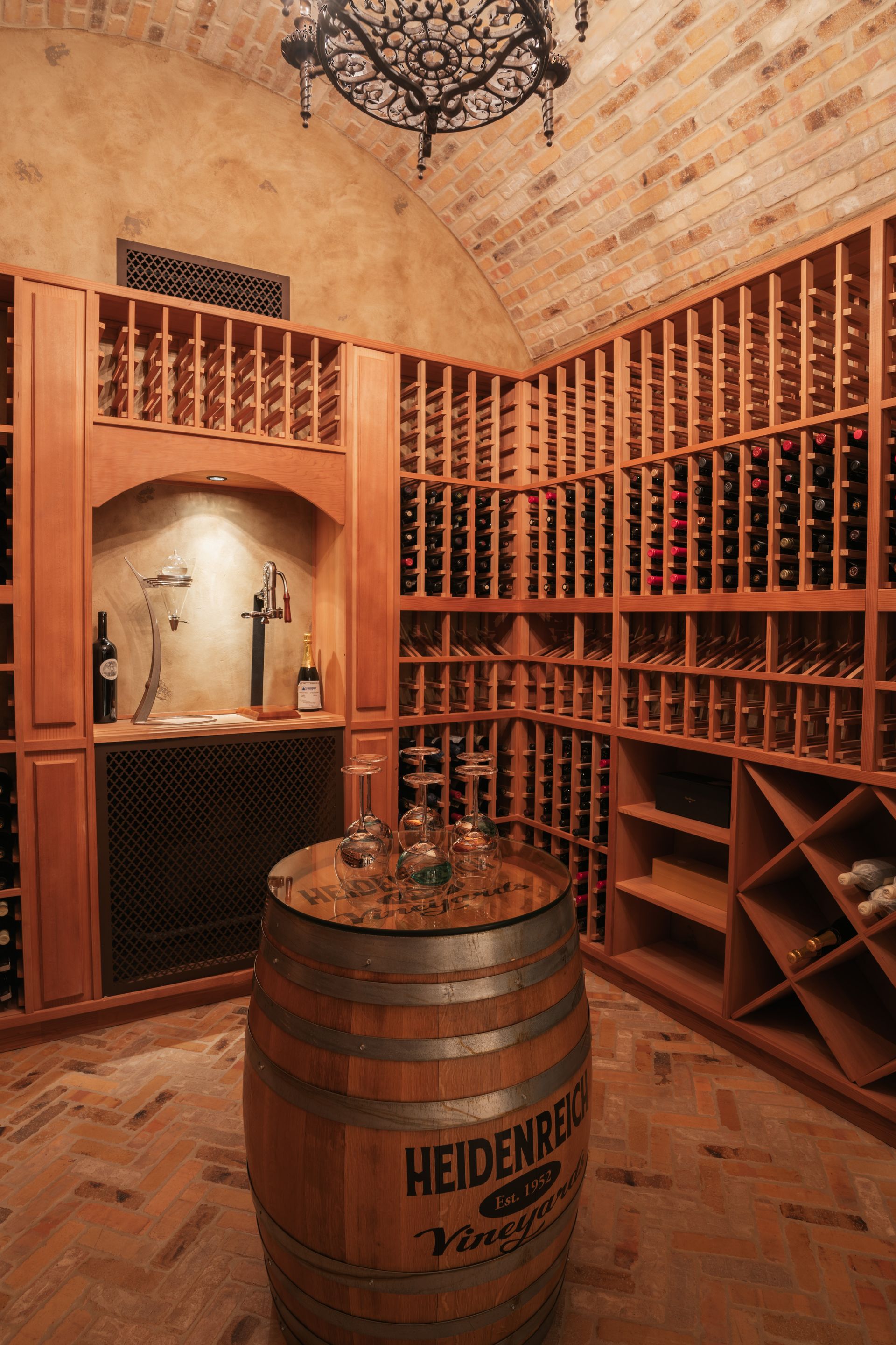 Wine cellar with wooden racks filled with bottles, arched brick ceiling, and a barrel table.