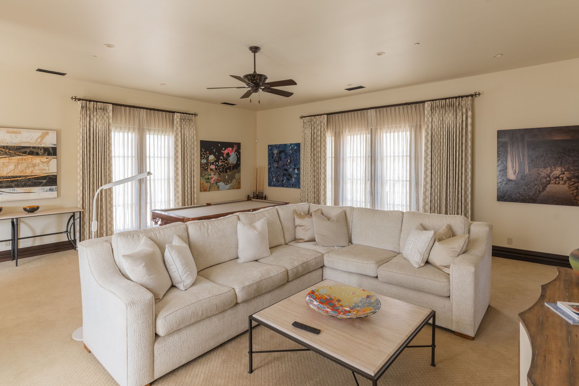 Living room with large cream sectional sofa, pool table, artwork, and windows with curtains.