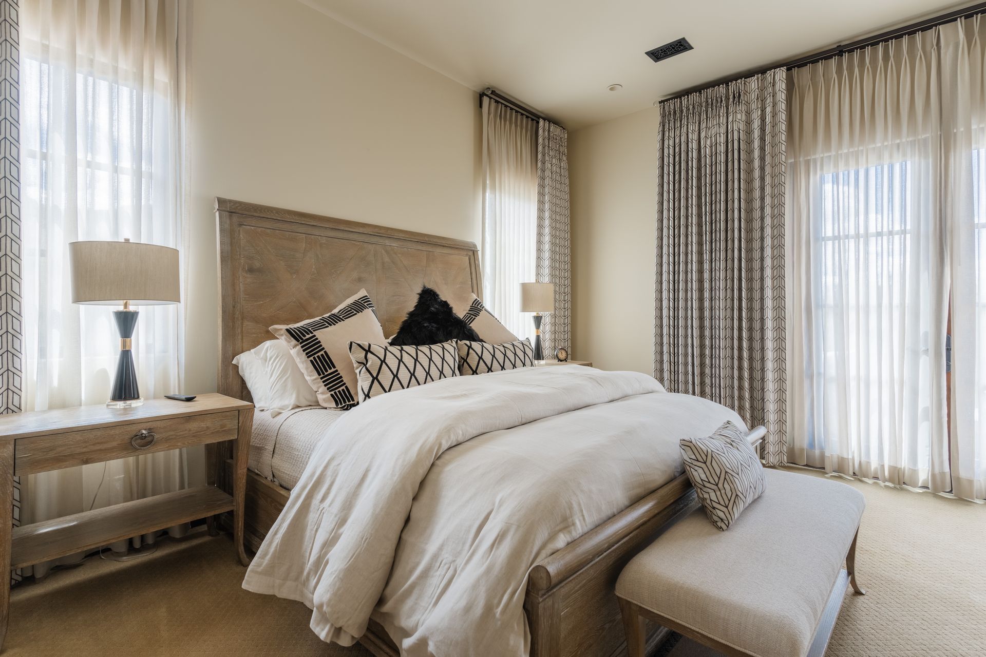 Bedroom with a bed, nightstands, and sheer curtains. Neutral tones with patterned pillows and curtains.