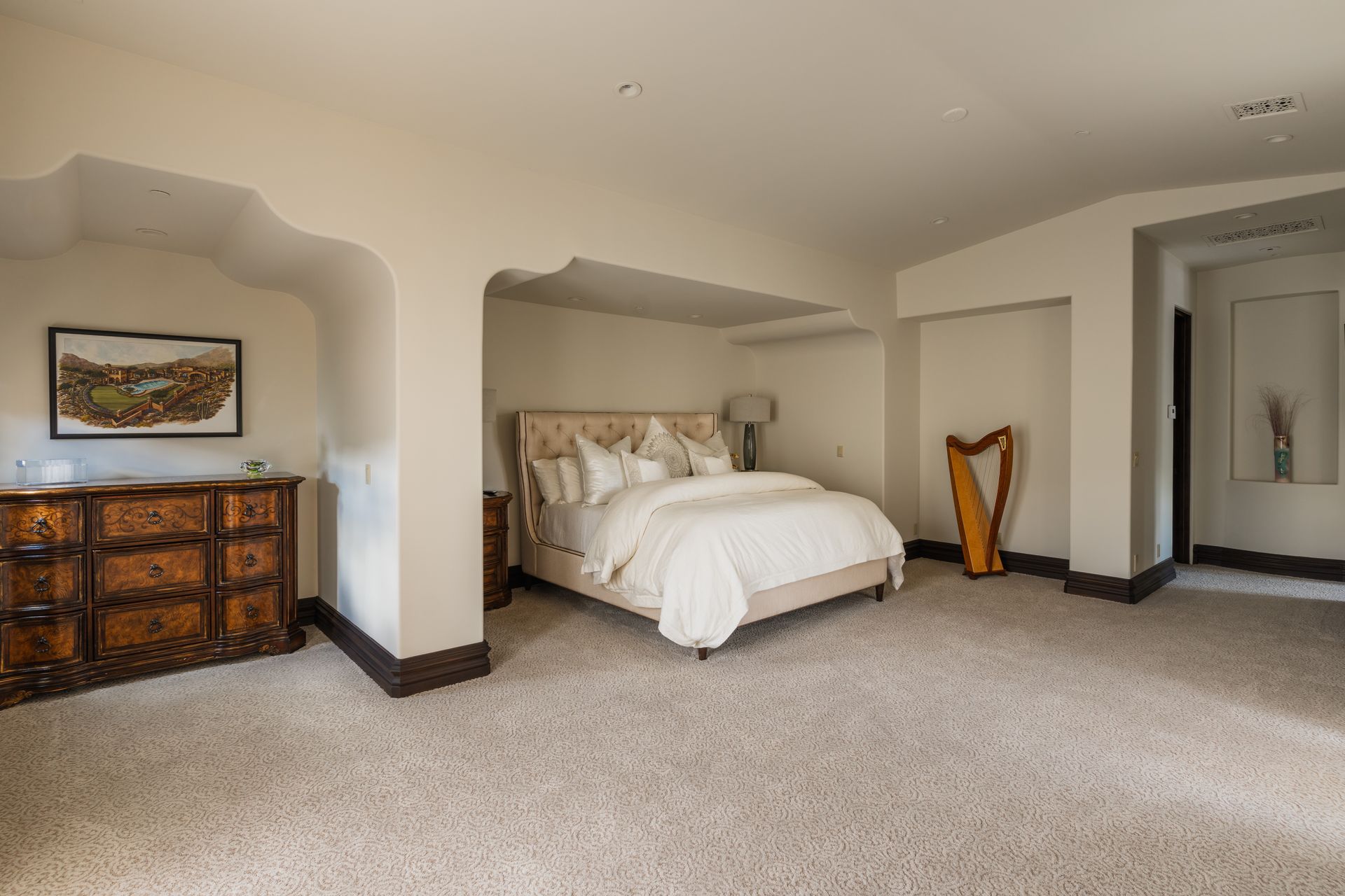 Spacious bedroom with bed, dresser, harp, and artwork on neutral carpet and walls.