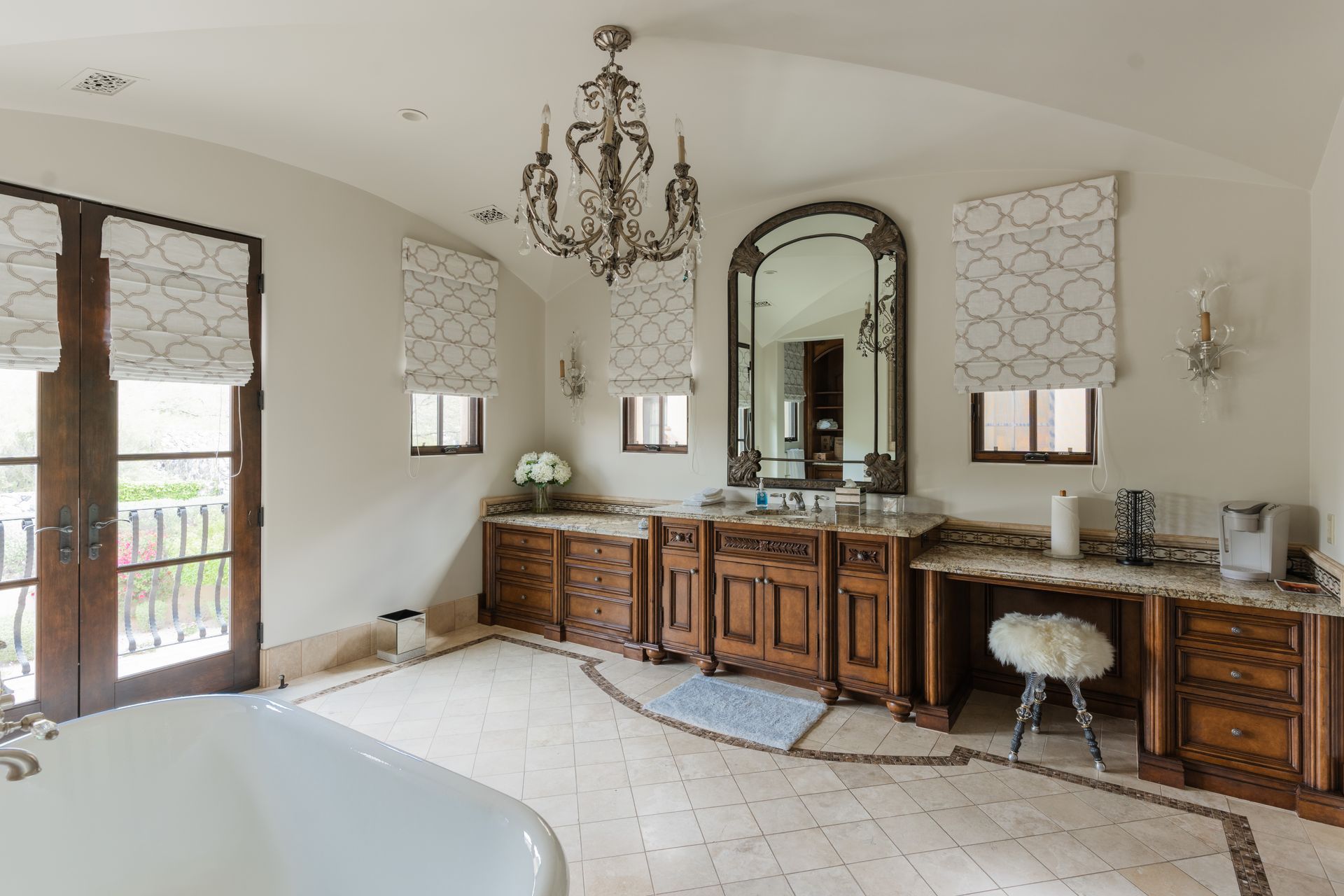 Elegant bathroom with a chandelier, wooden cabinets, arched mirror, and patterned Roman shades.