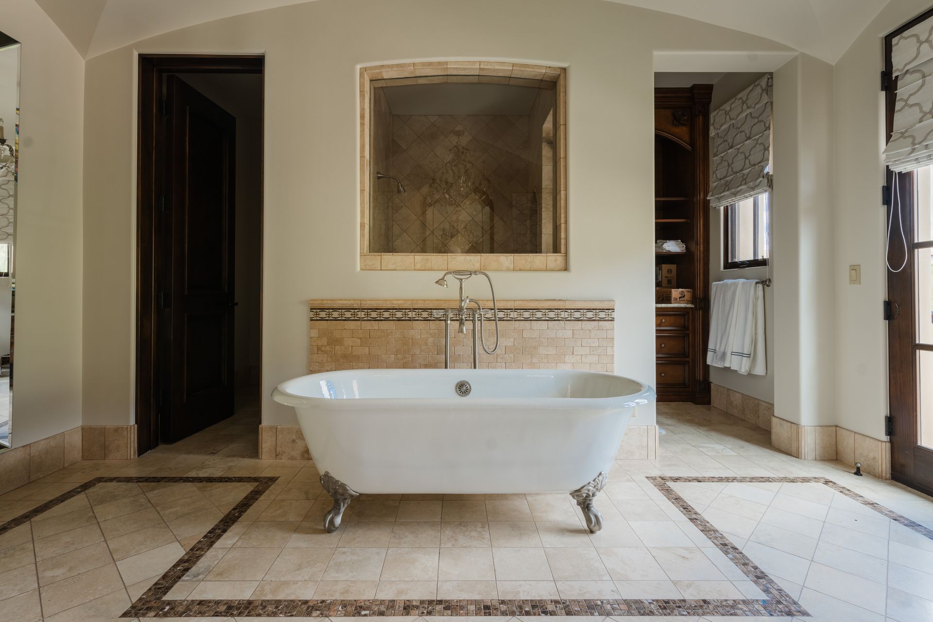 Clawfoot bathtub in a luxurious bathroom with neutral-toned tiling, a large mirror, and dark wooden doors.