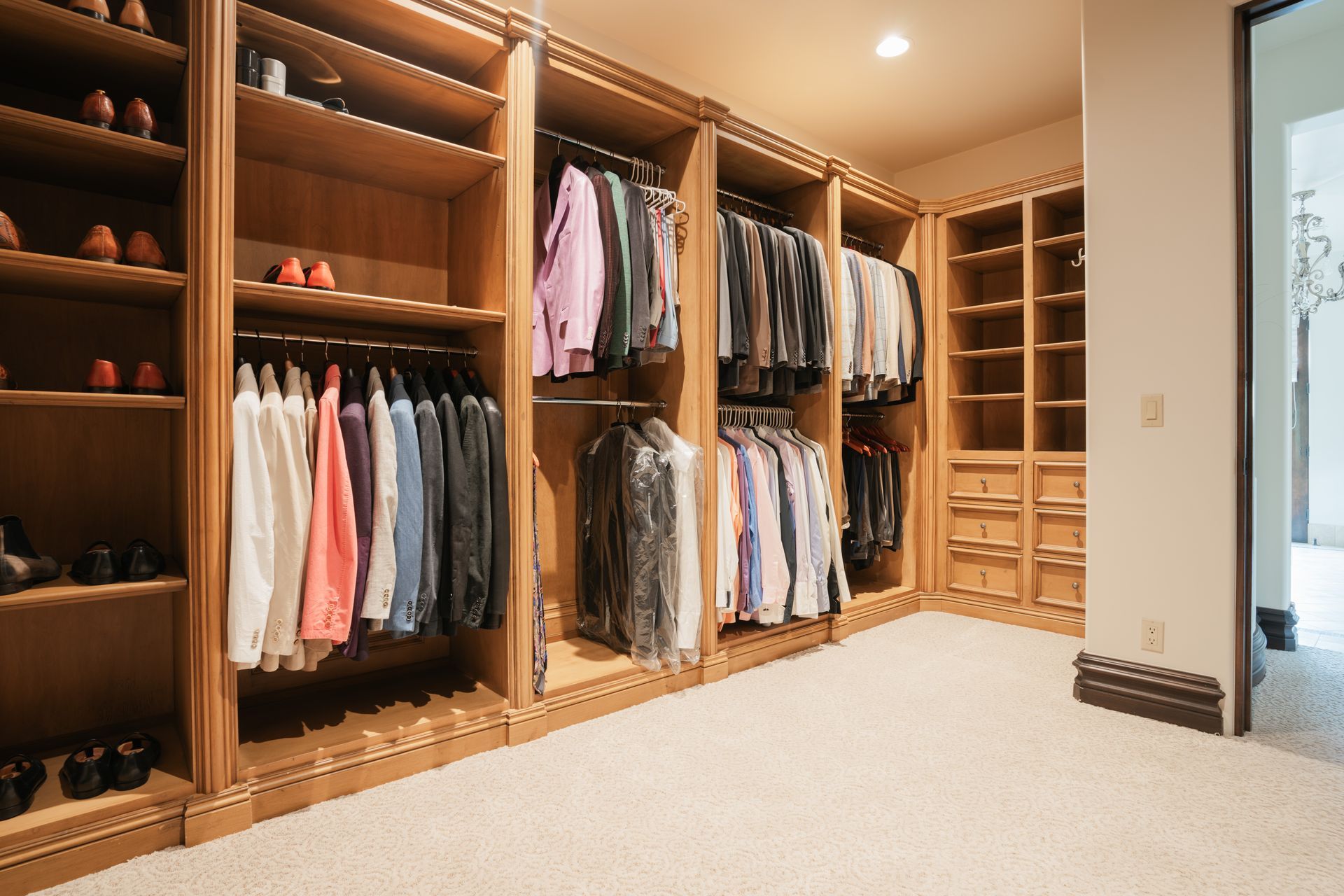 Walk-in closet with light wood shelving and hanging clothes, beige carpet, and a white wall.