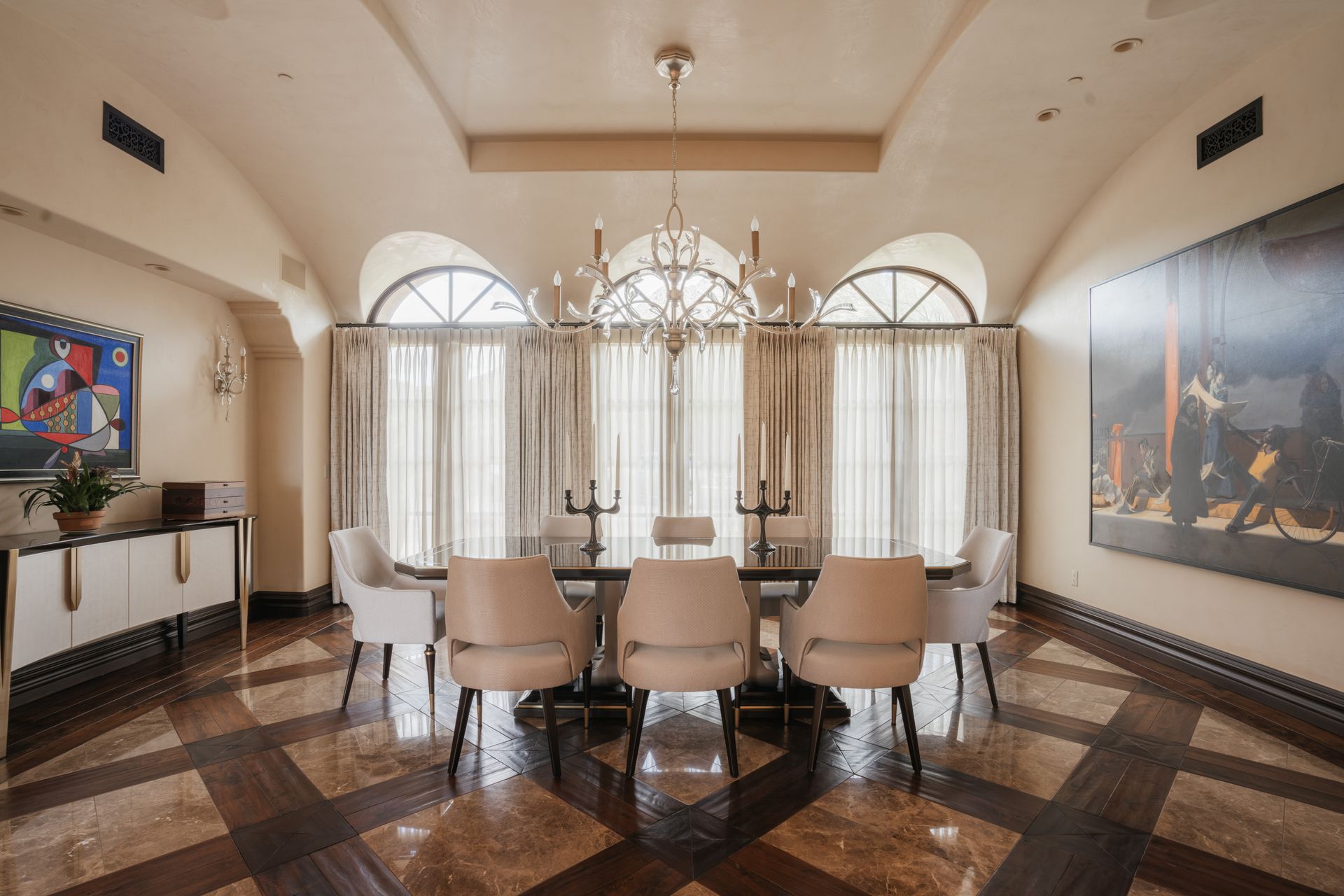 Elegant dining room with arched windows, chandelier, long table, chairs, art, and patterned floor.