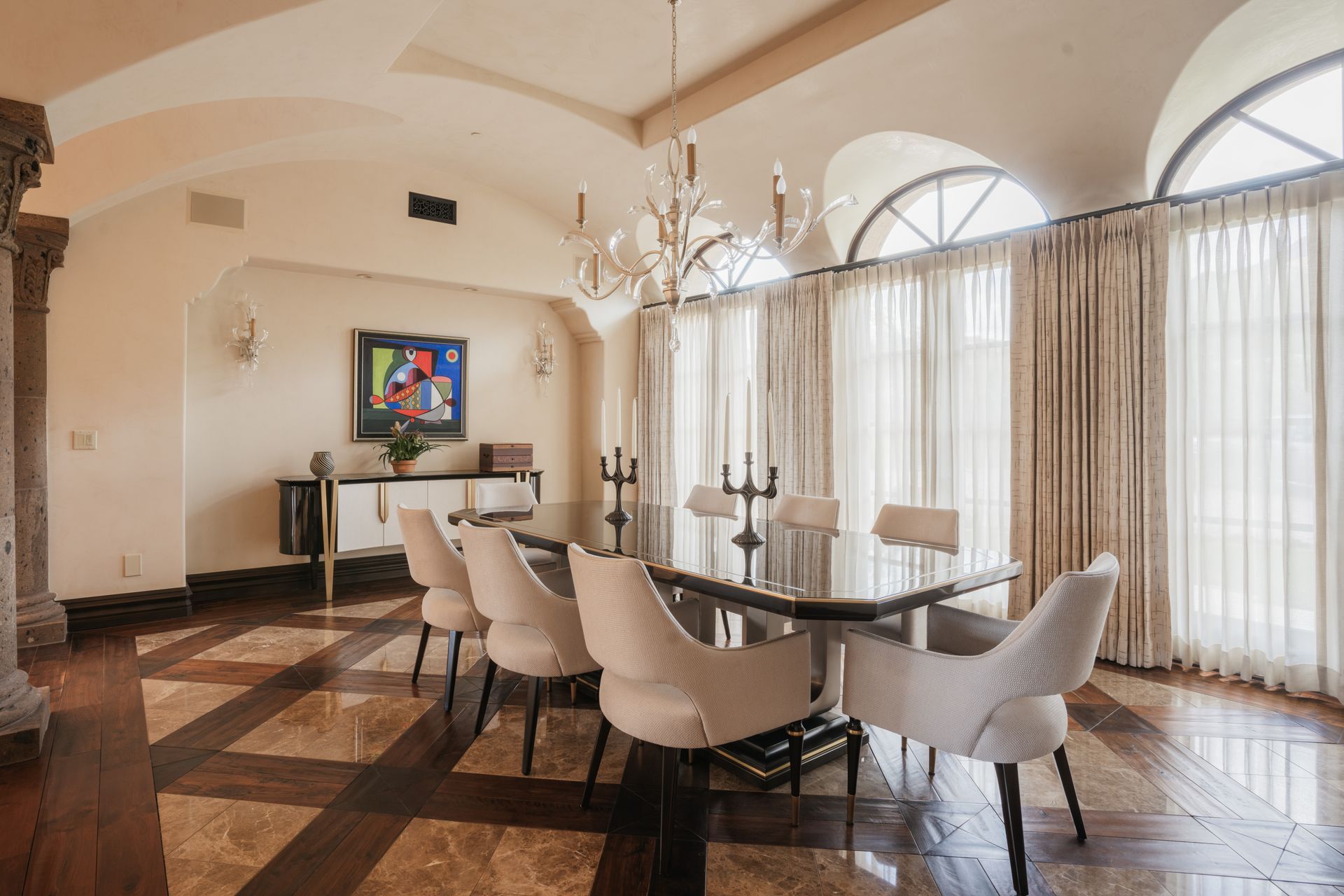 Elegant dining room with chandelier, dark table, white chairs, arched windows, and patterned floor.