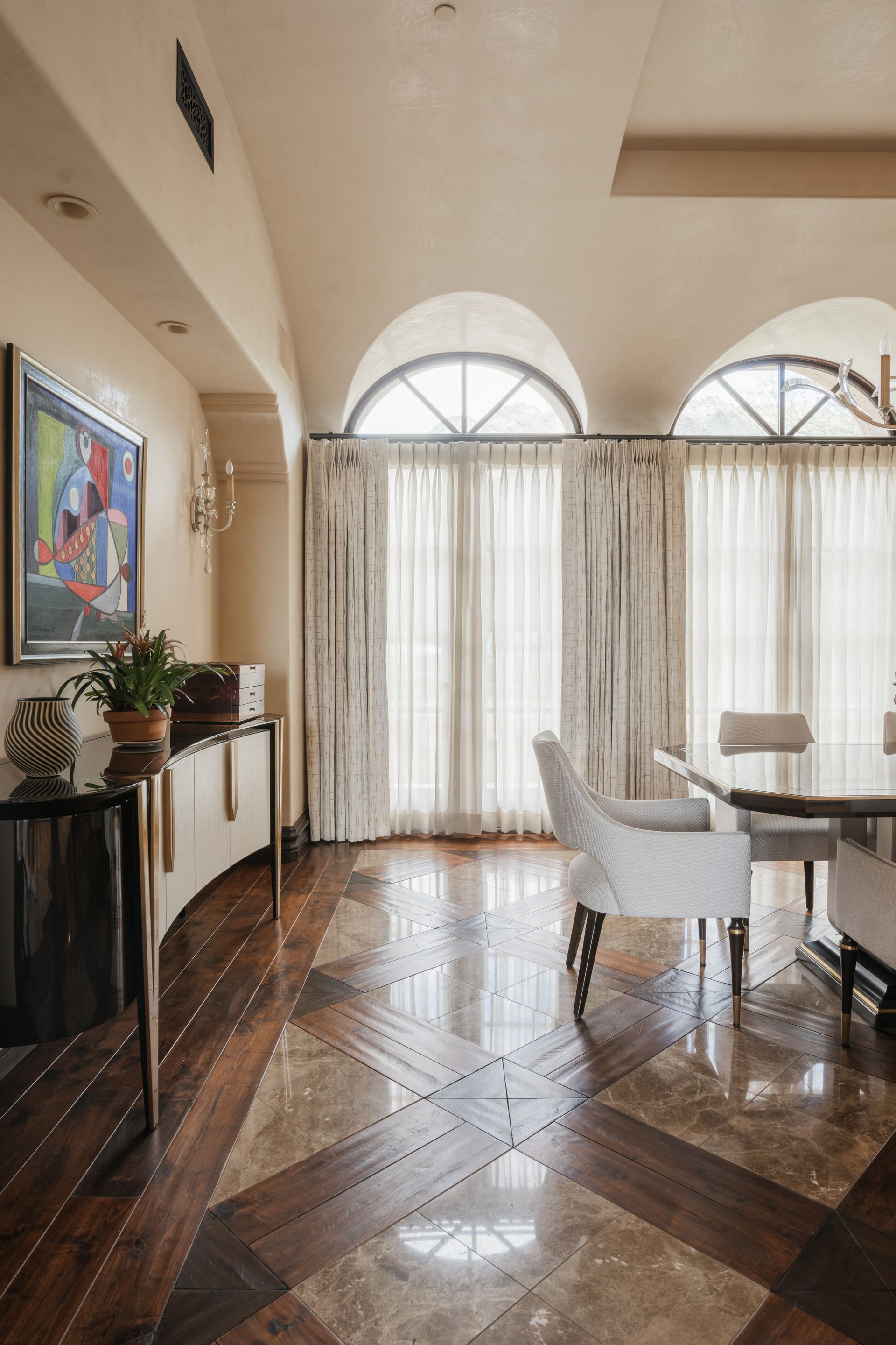 Elegant dining room with arched windows, light-colored walls, patterned wood floors, and a modern buffet.