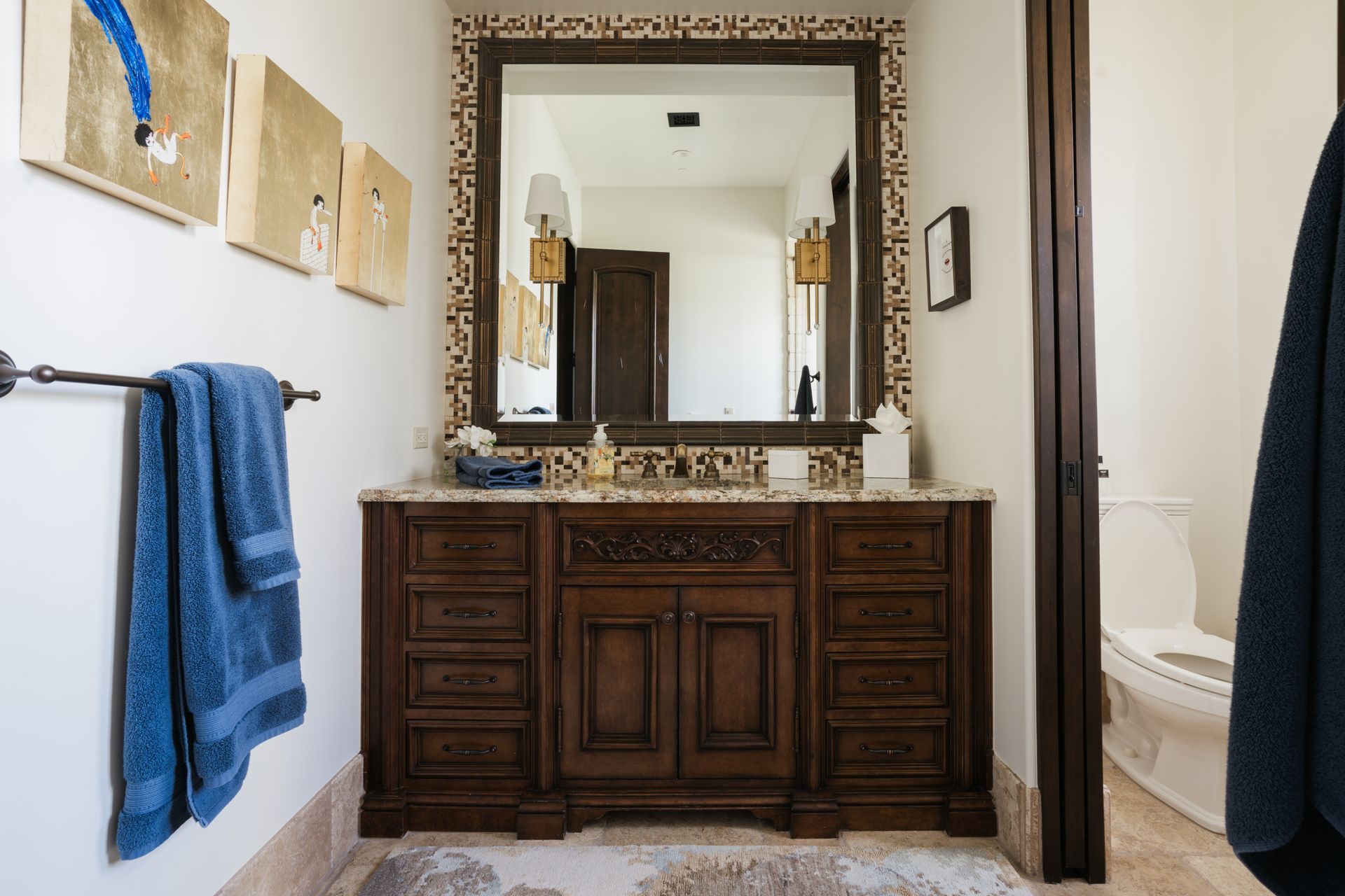 Bathroom with dark wood vanity, mirror with mosaic frame, blue towels, and artwork.