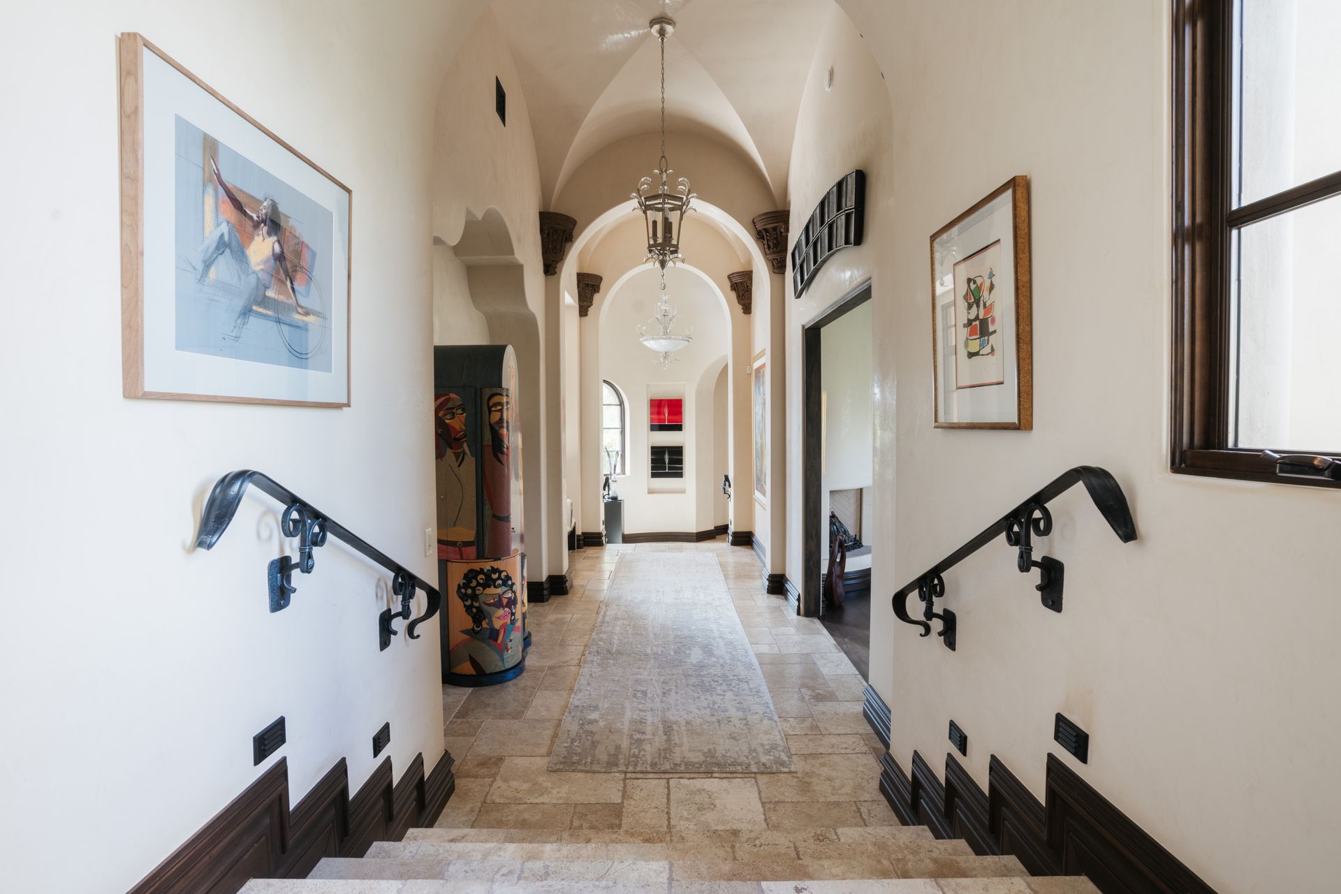 Hallway with arched ceiling, artwork, stairs with ornate railing, and a patterned stone floor.