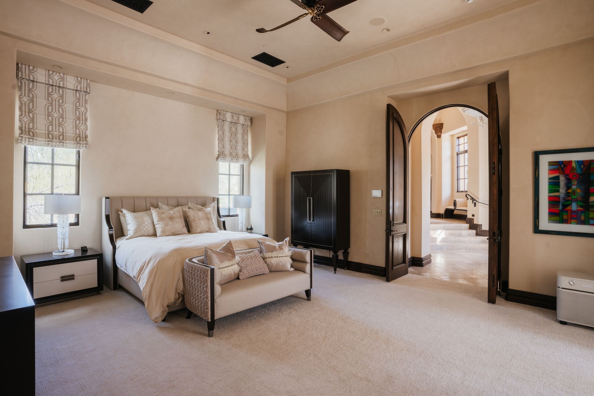 Spacious bedroom with bed, wardrobe, bench, and arched doorway to a hallway. Neutral colors, natural light.