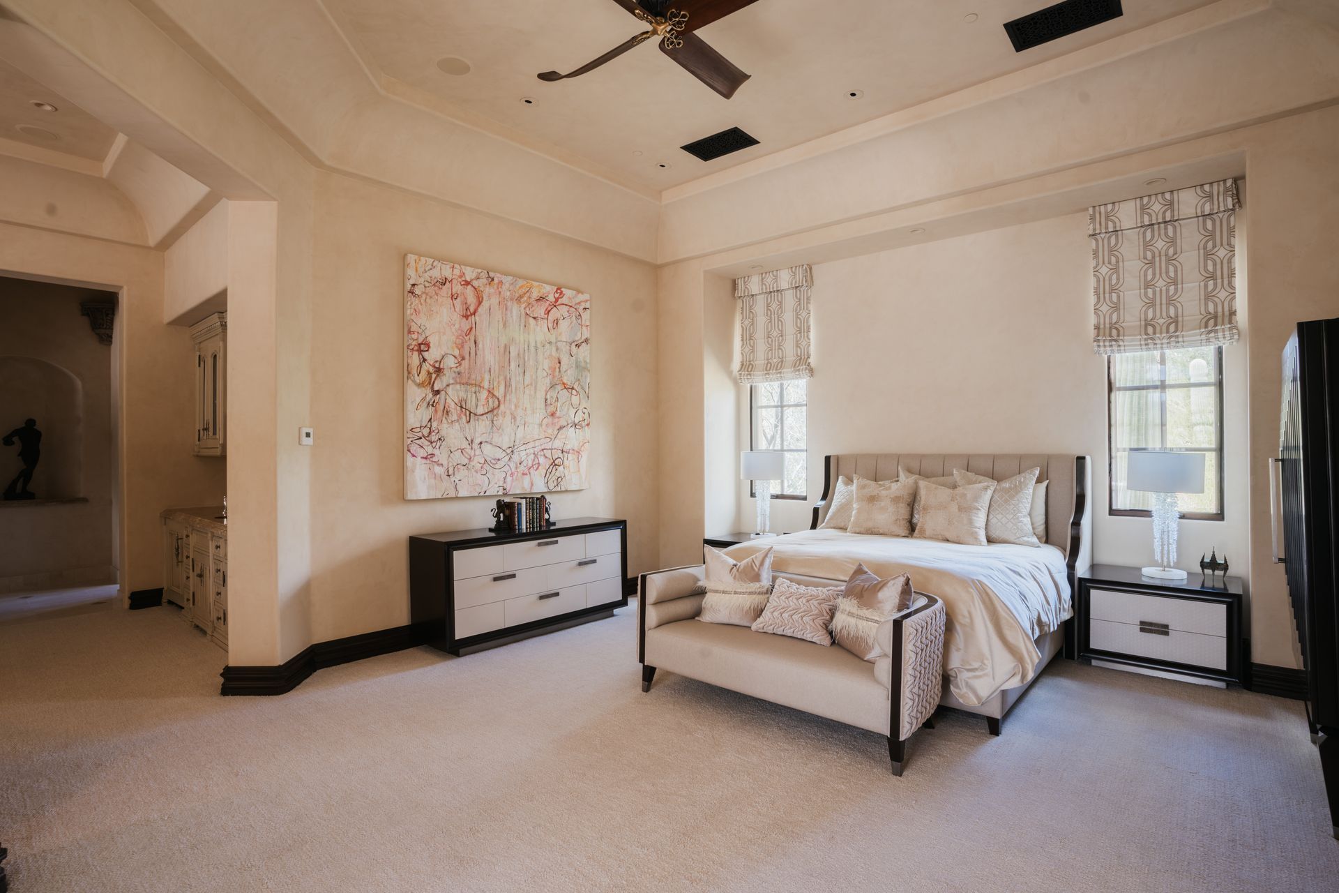 Spacious bedroom with cream walls, carpet, and a large bed, featuring a dresser, artwork, and windows.