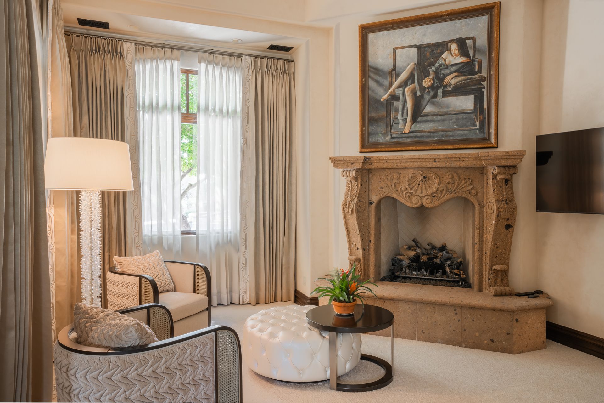 Living room with fireplace, armchairs, and round ottoman in front of a window with sheer curtains.