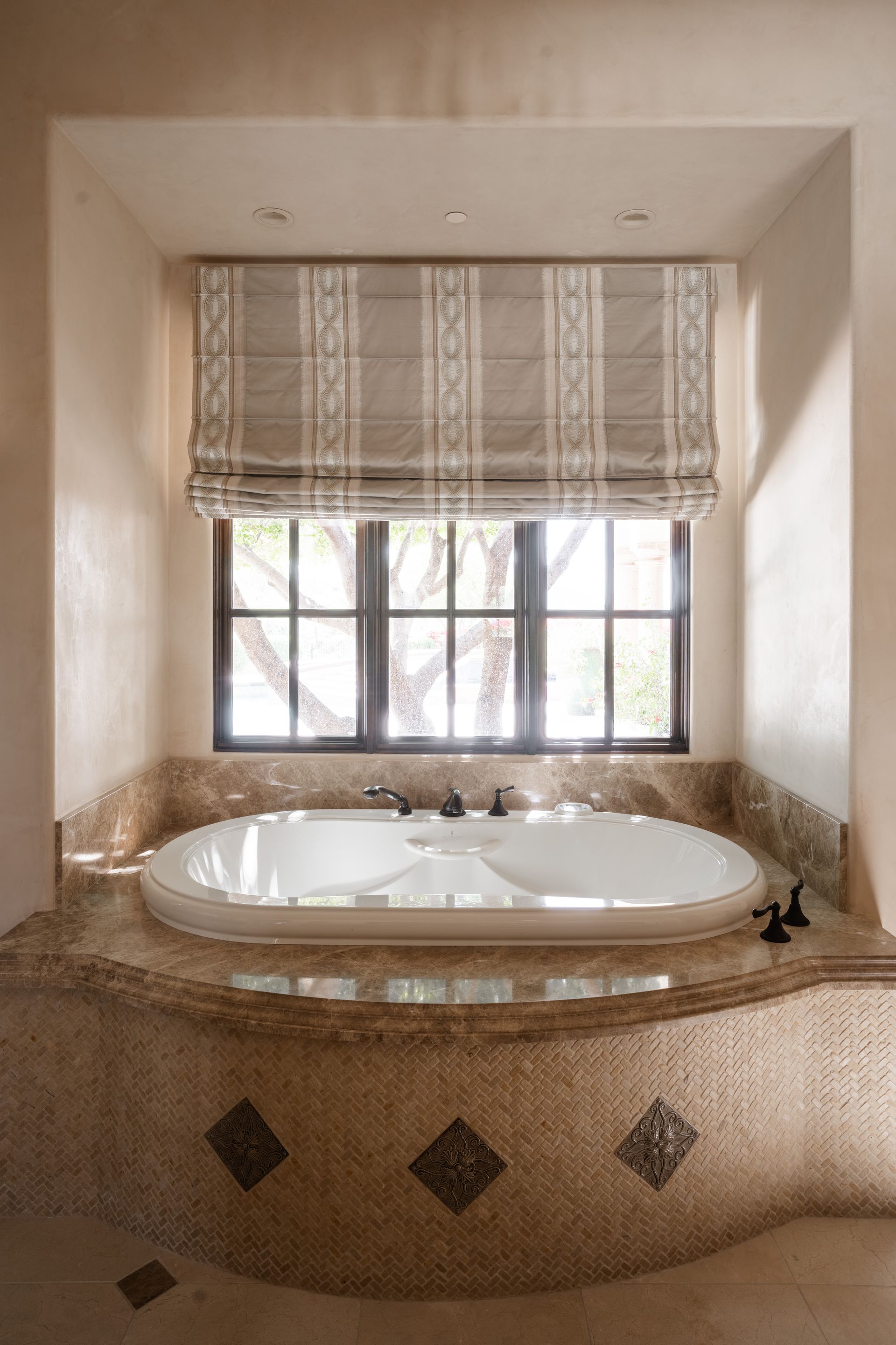 Bathtub alcove with mosaic tile, oval tub, and Roman shade window treatment.