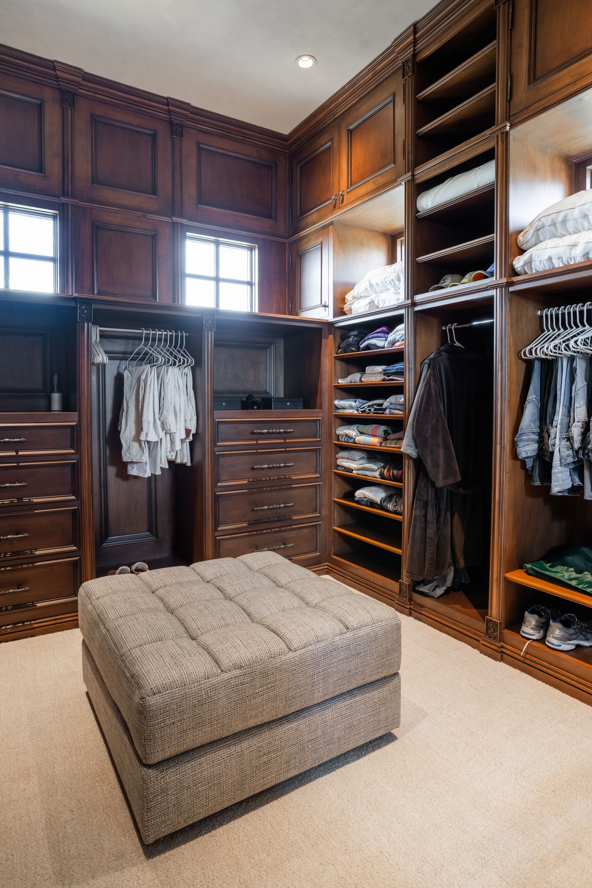 Walk-in closet with dark wood shelving, drawers, and hanging rods. Includes an upholstered ottoman and carpeted floor.