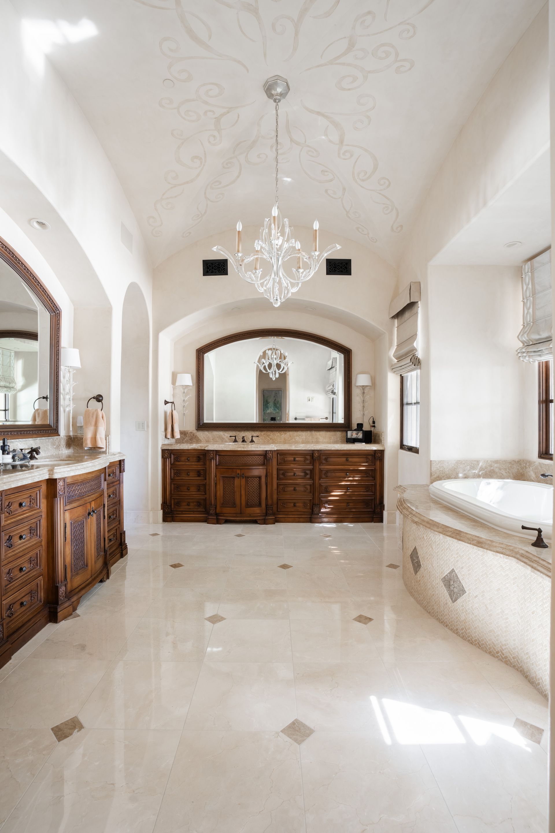 Luxurious bathroom with carved wood vanities, marble floor, chandelier, and a mosaic-tiled tub.