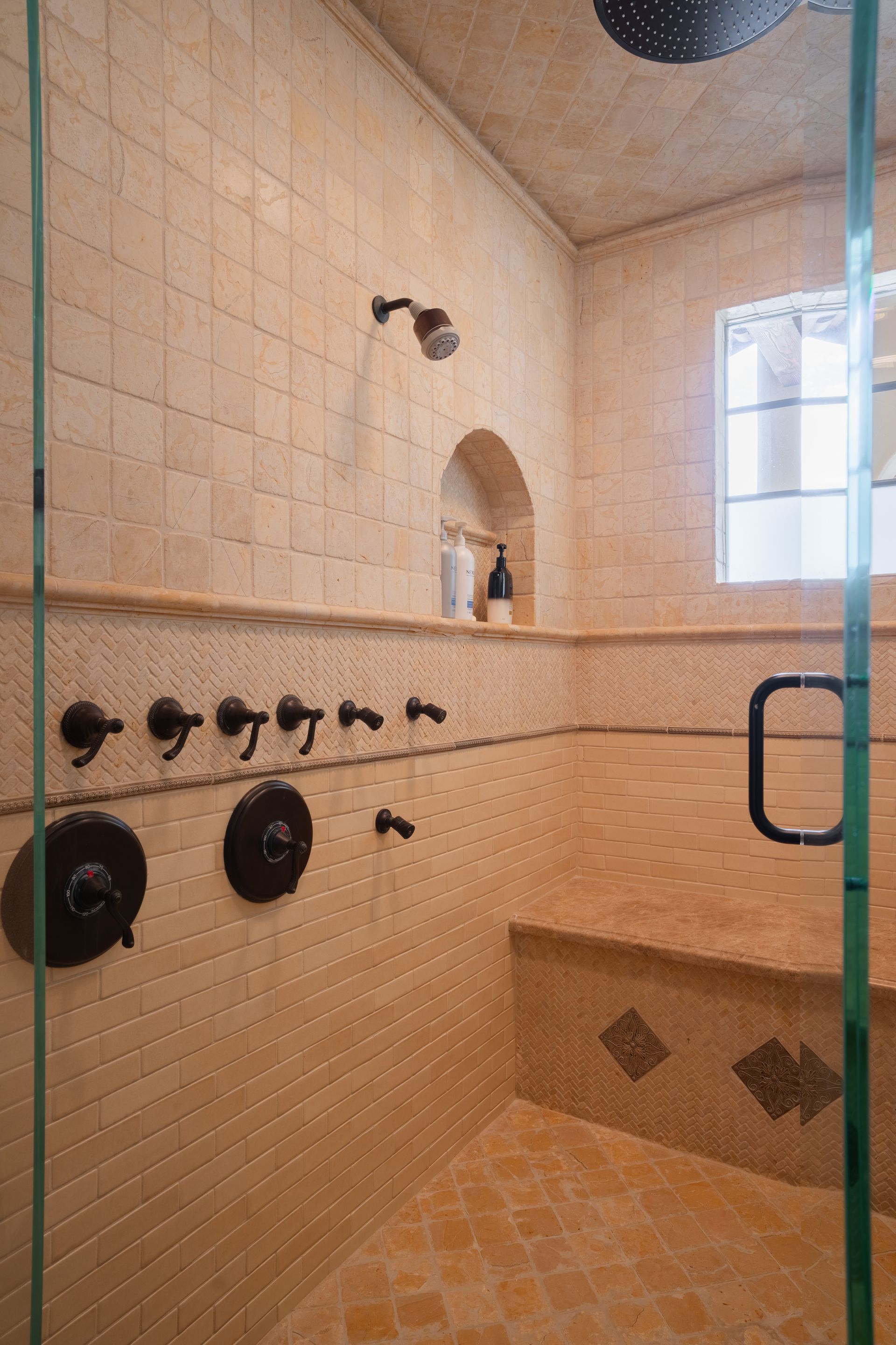 Shower with beige tile walls, multiple shower heads, and glass door.