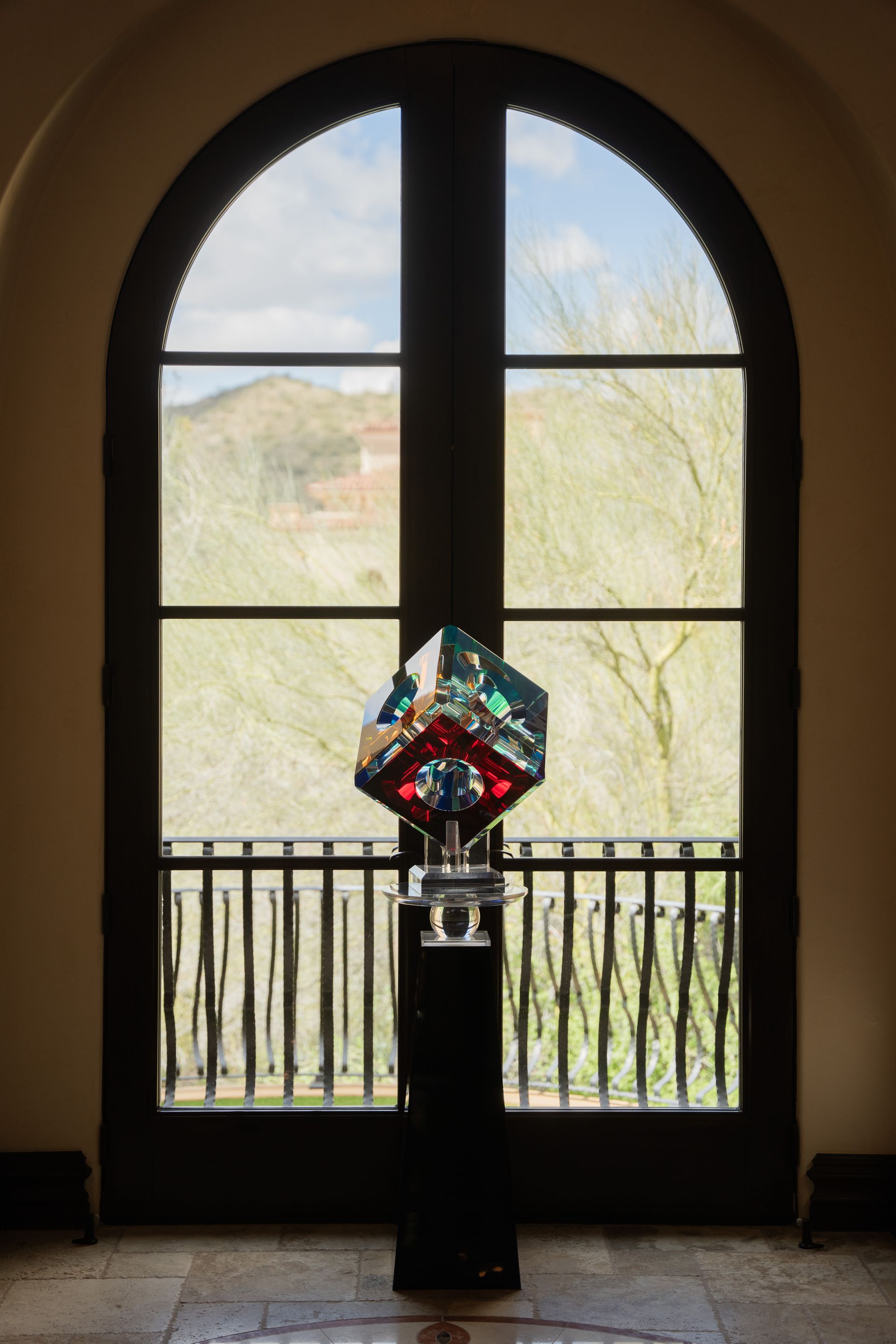 A colorful cube sculpture on a black pedestal, in front of an arched window overlooking a balcony.