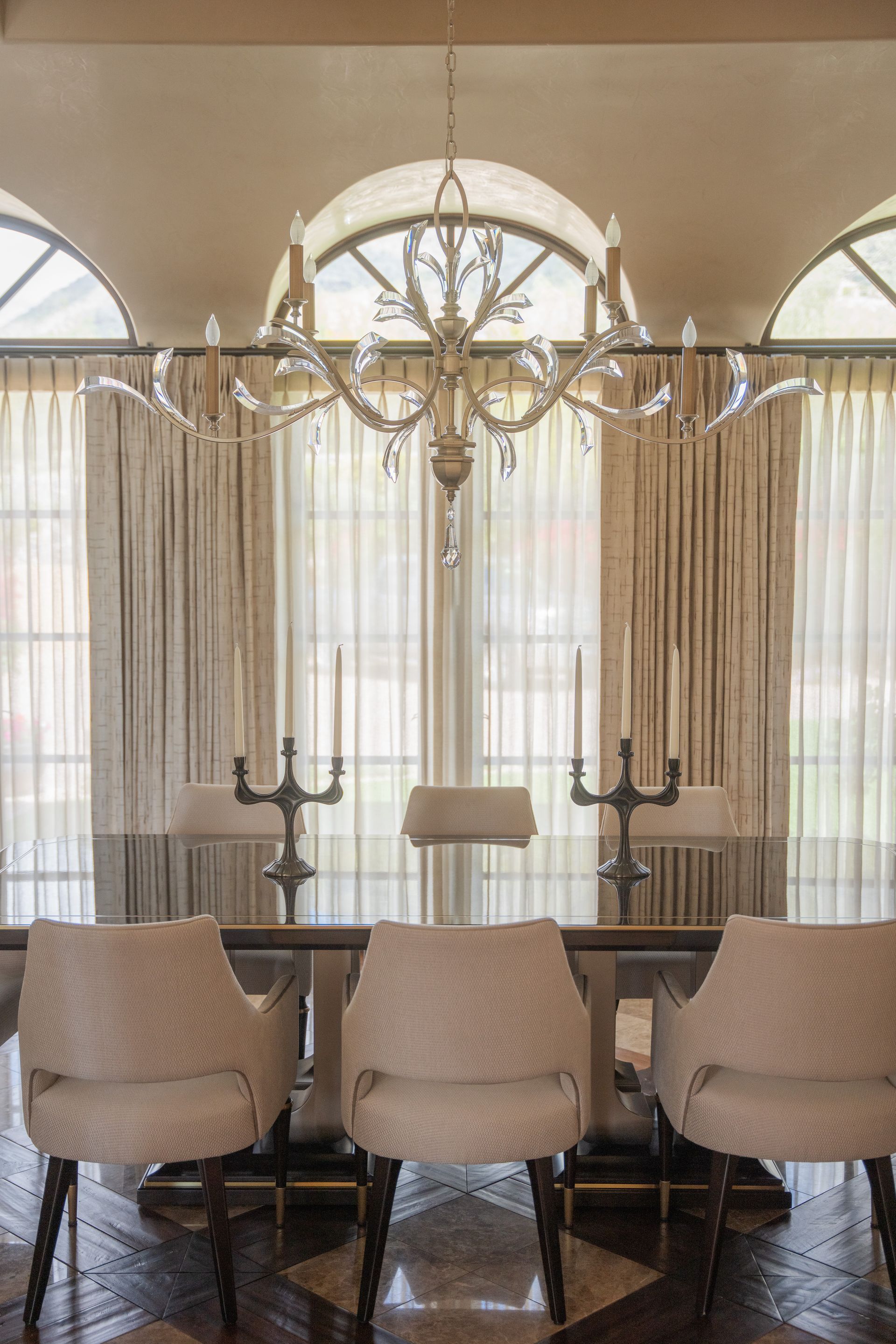 Dining room with a chandelier above a table with three chairs, arched windows, and cream curtains.