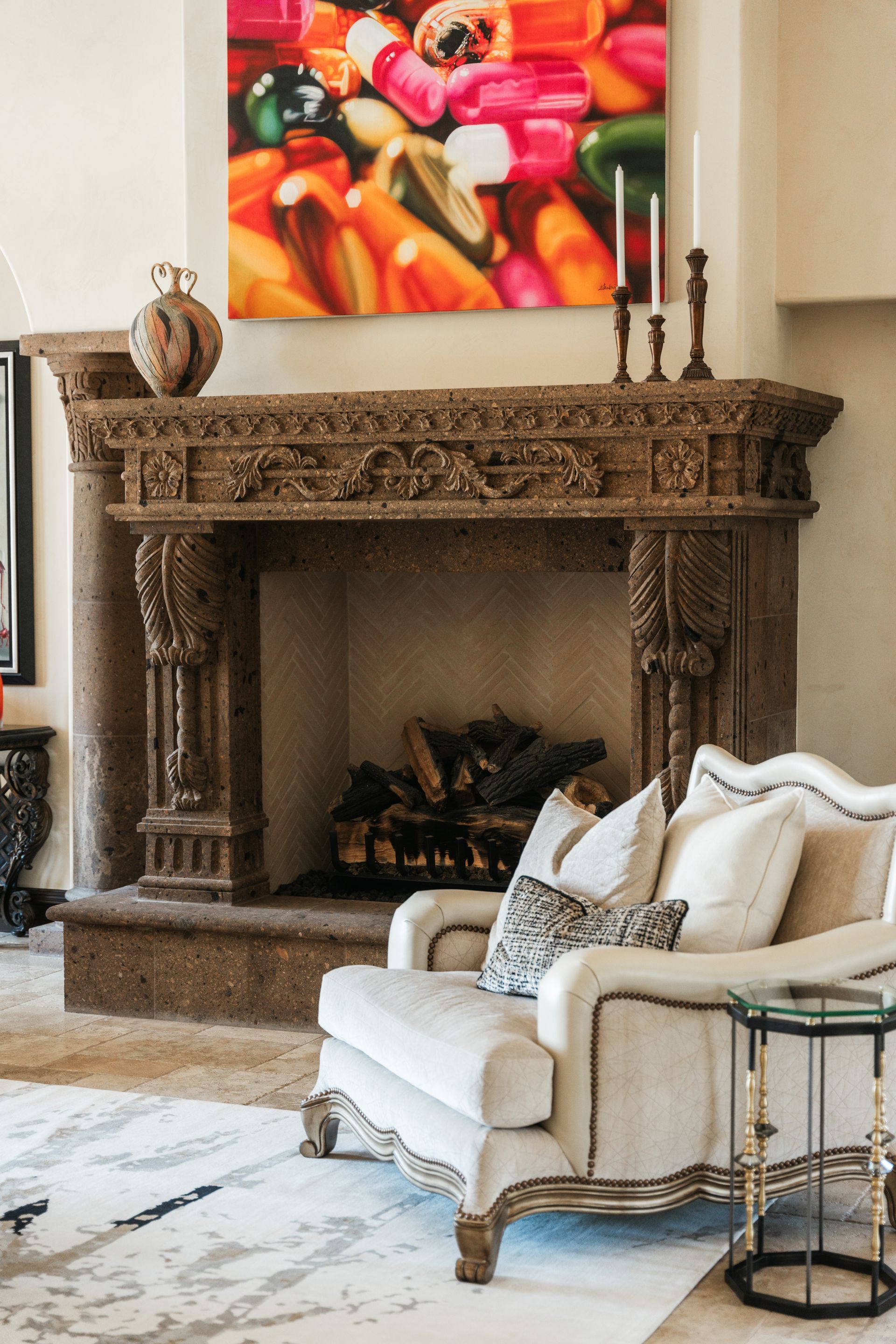 Ornate stone fireplace with carved details, colorful painting above, and upholstered armchair.